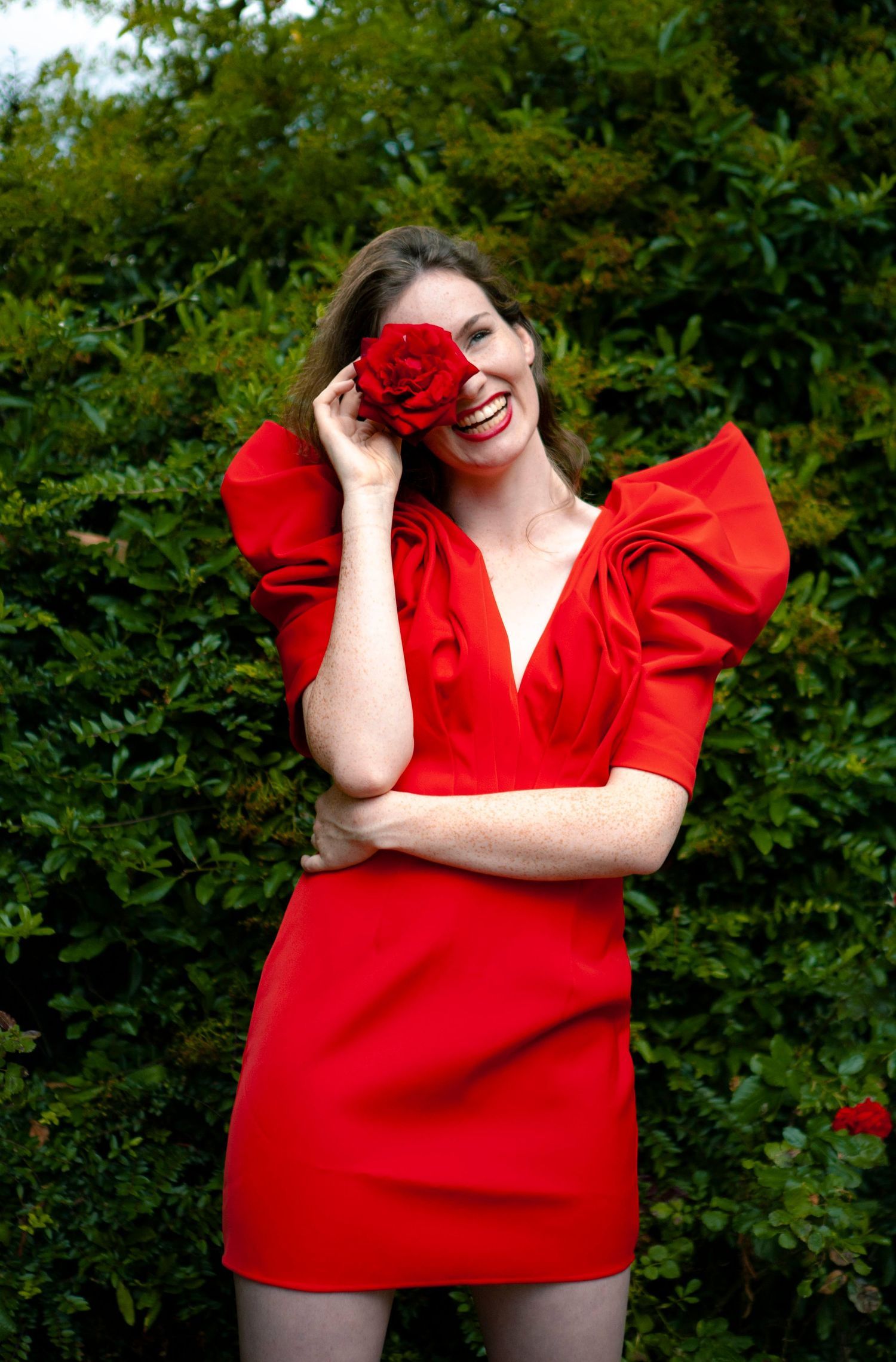 Fashion portrait in a vibrant red dress holding a rose against green foliage background.