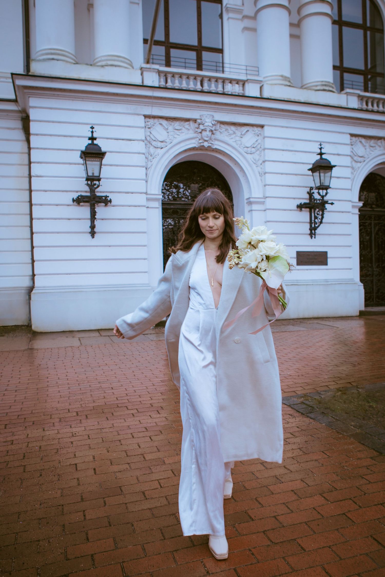 A person in a stylish white pantsuit and long coat holds a bouquet while walking on brick pavement outside an elegant building.