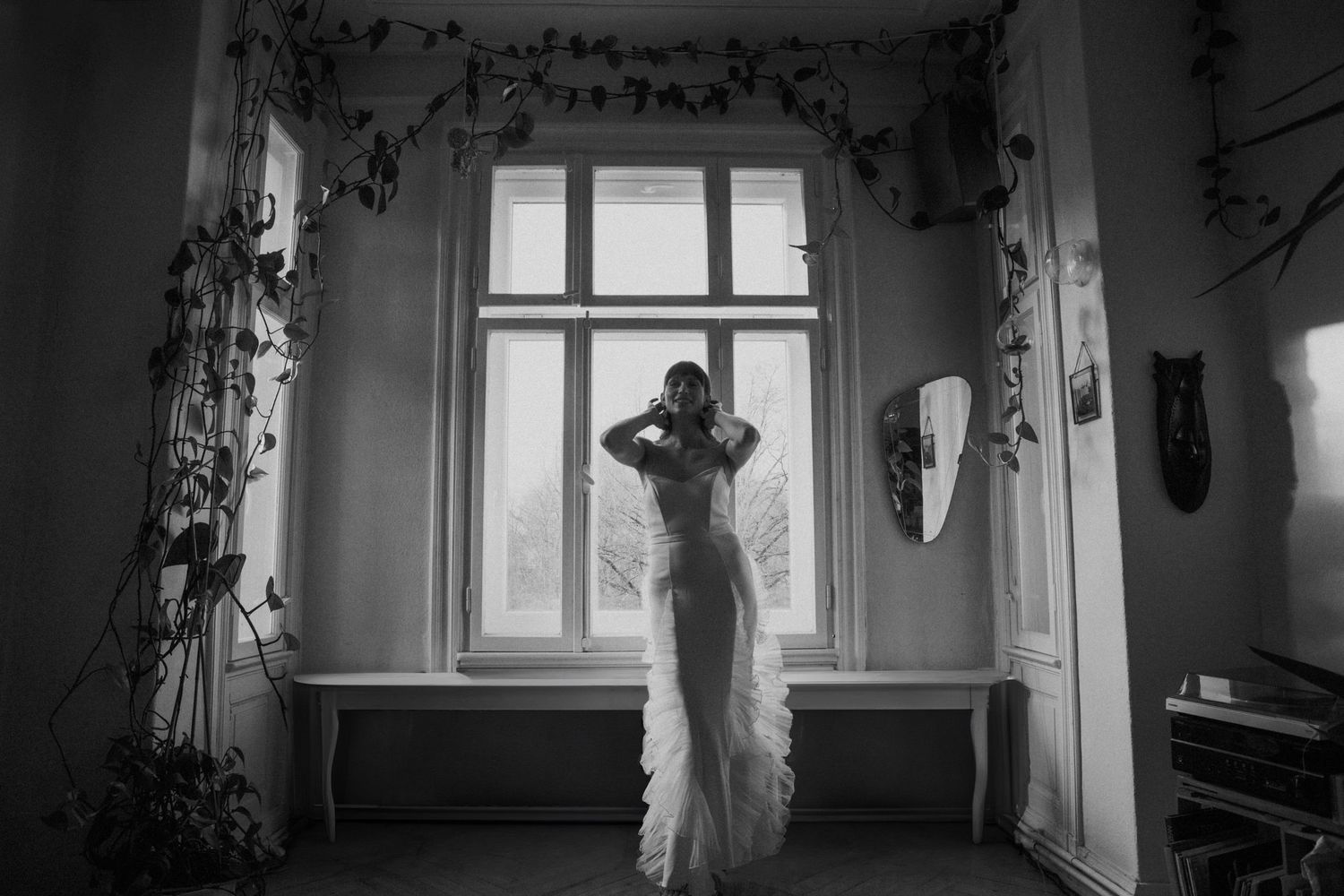 Black and white photo of silhouette in flowing wedding dress standing by ornate window.