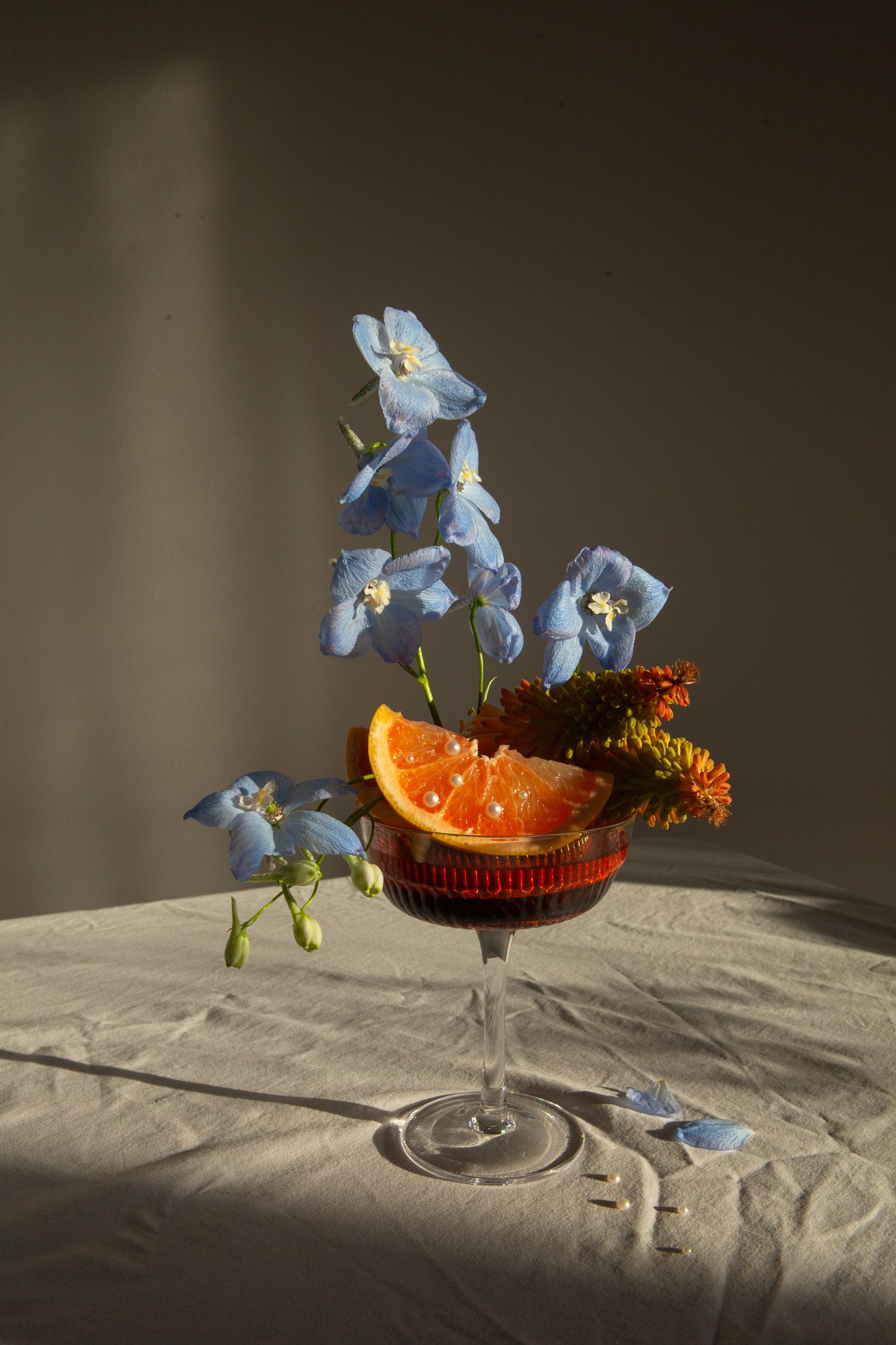 Blue delphinium flowers and orange slices arranged in a cocktail glass with dramatic lighting.