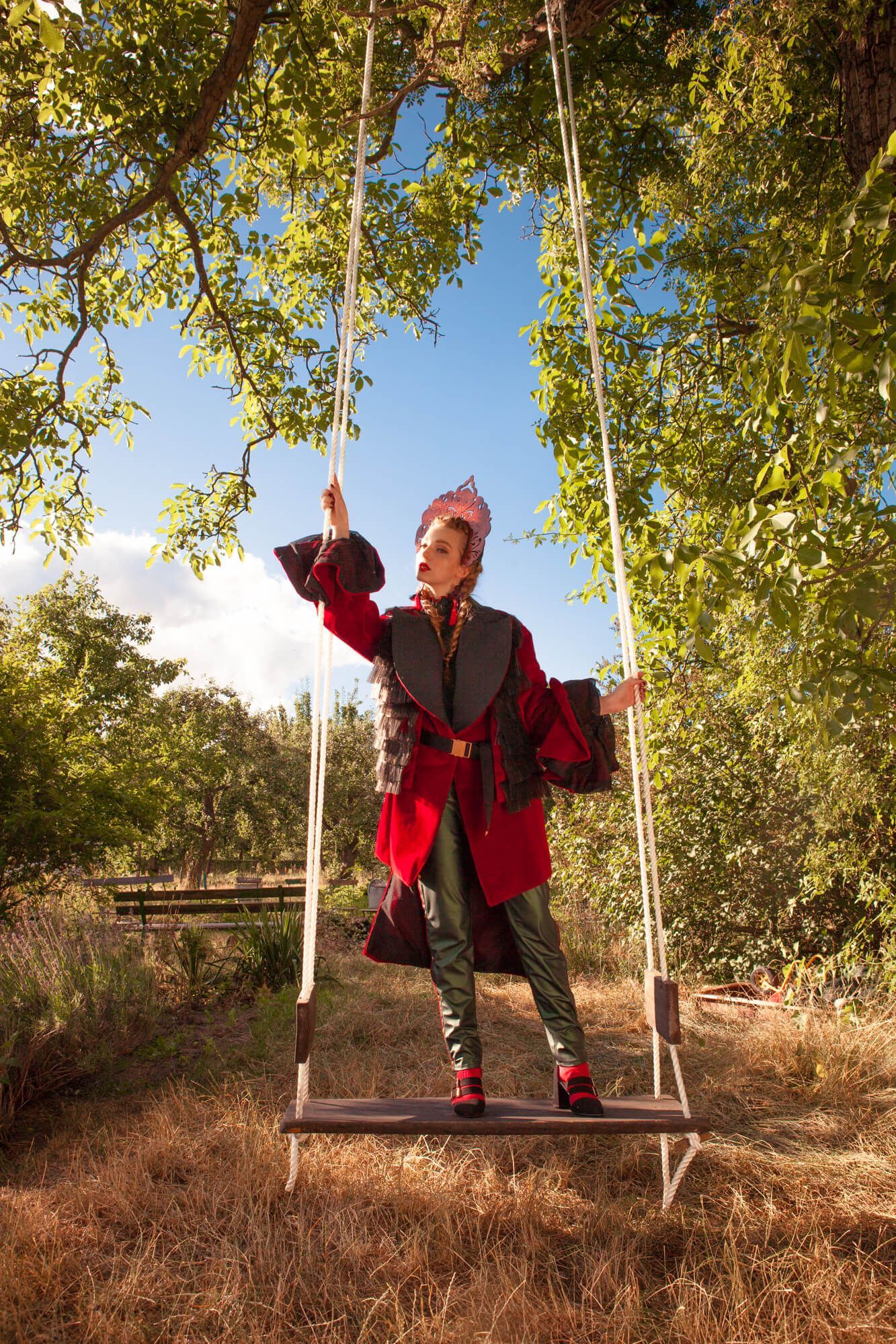 Figure in a red vintage coat stands balanced on a rustic wooden swing in an autumn garden setting.
