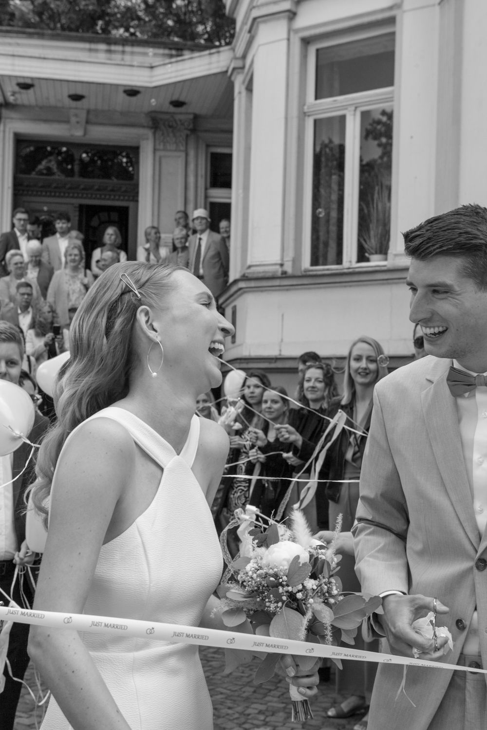 A black and white wedding photograph showing a joyful moment between newlyweds surrounded by guests outside a building.