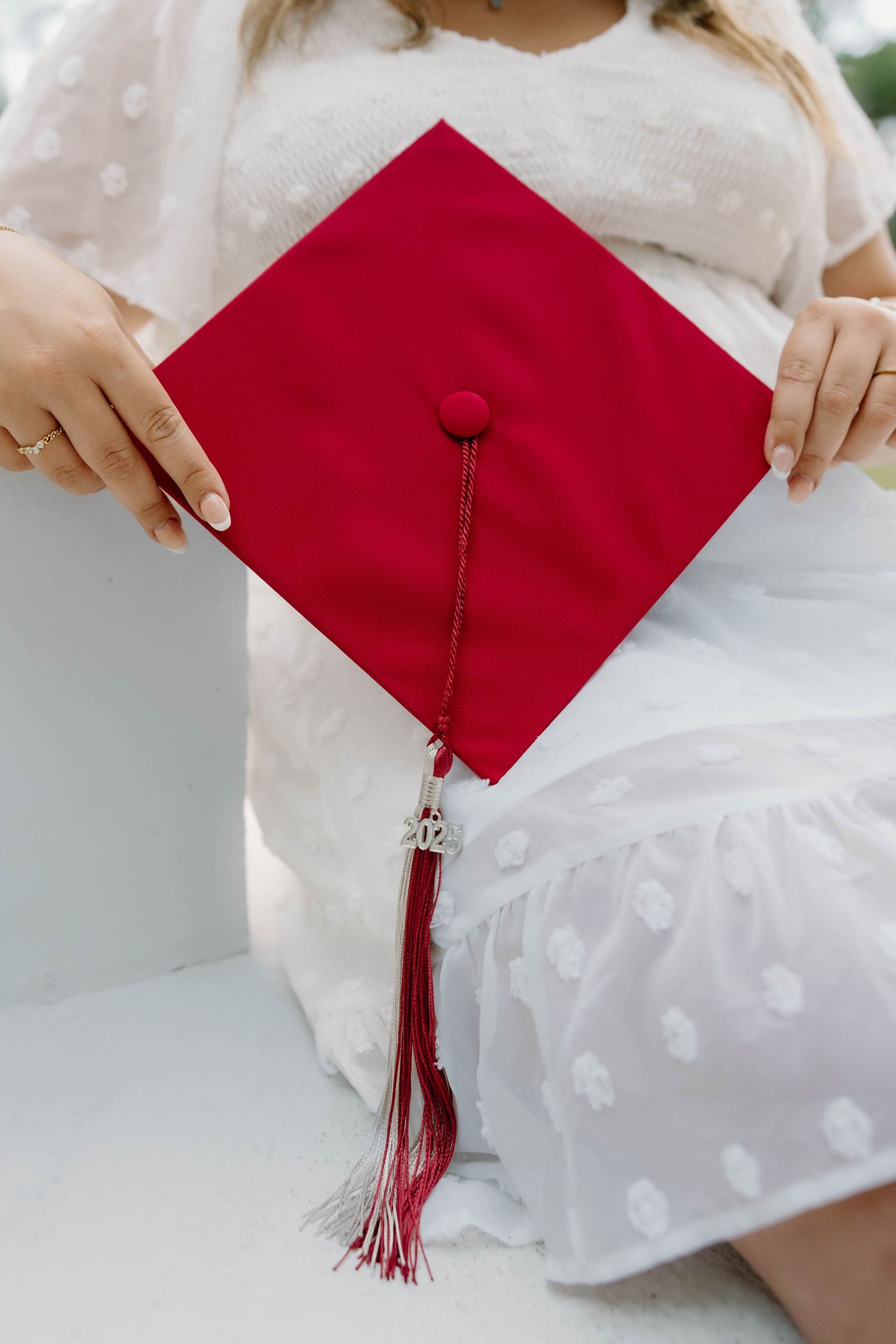 A graduate in a white dress holds a red mortarboard graduation cap with tassel and year charm.