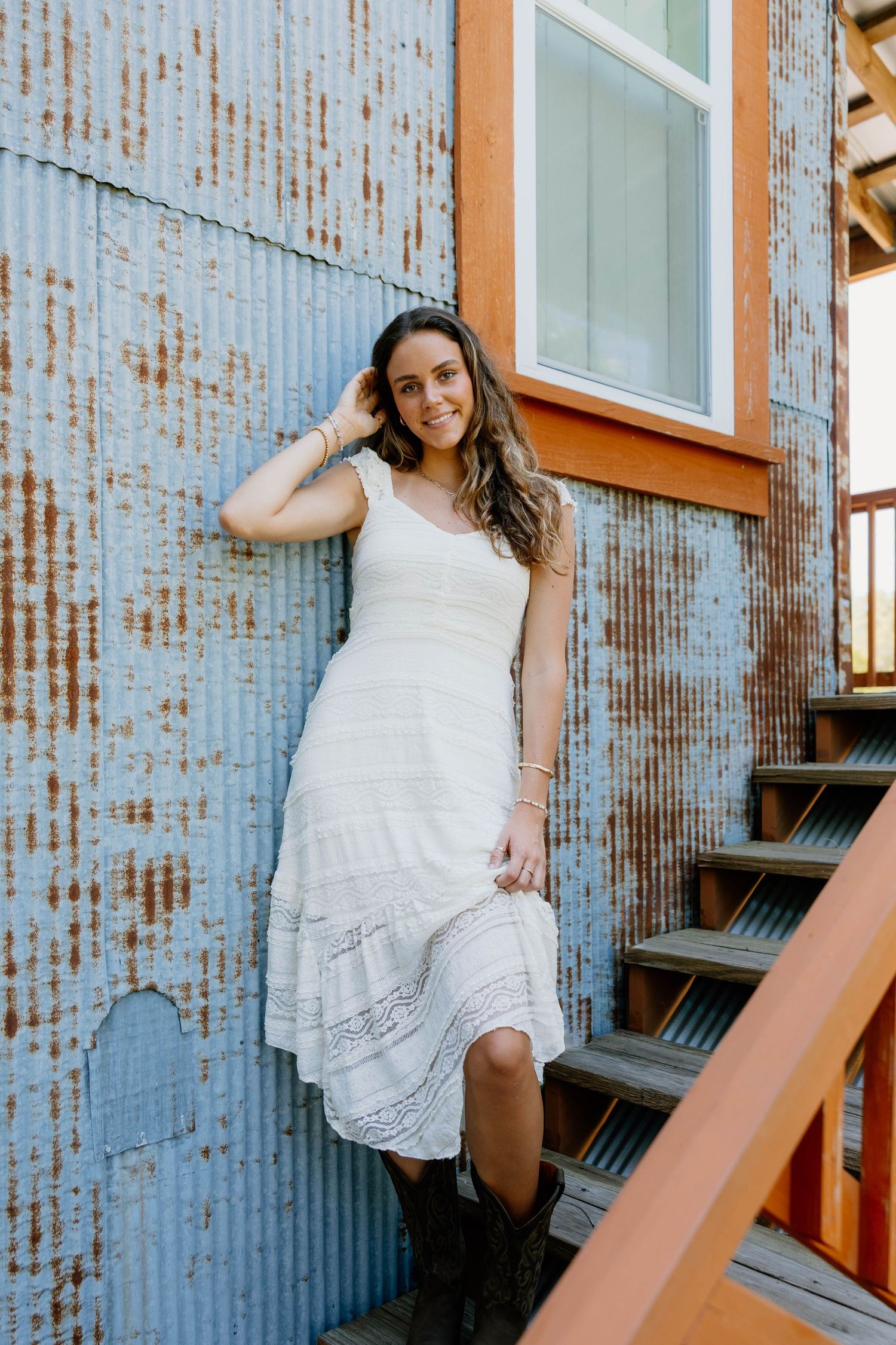 Woman in white midi dress and boots leaning against rustic corrugated metal wall near wooden stairs.
