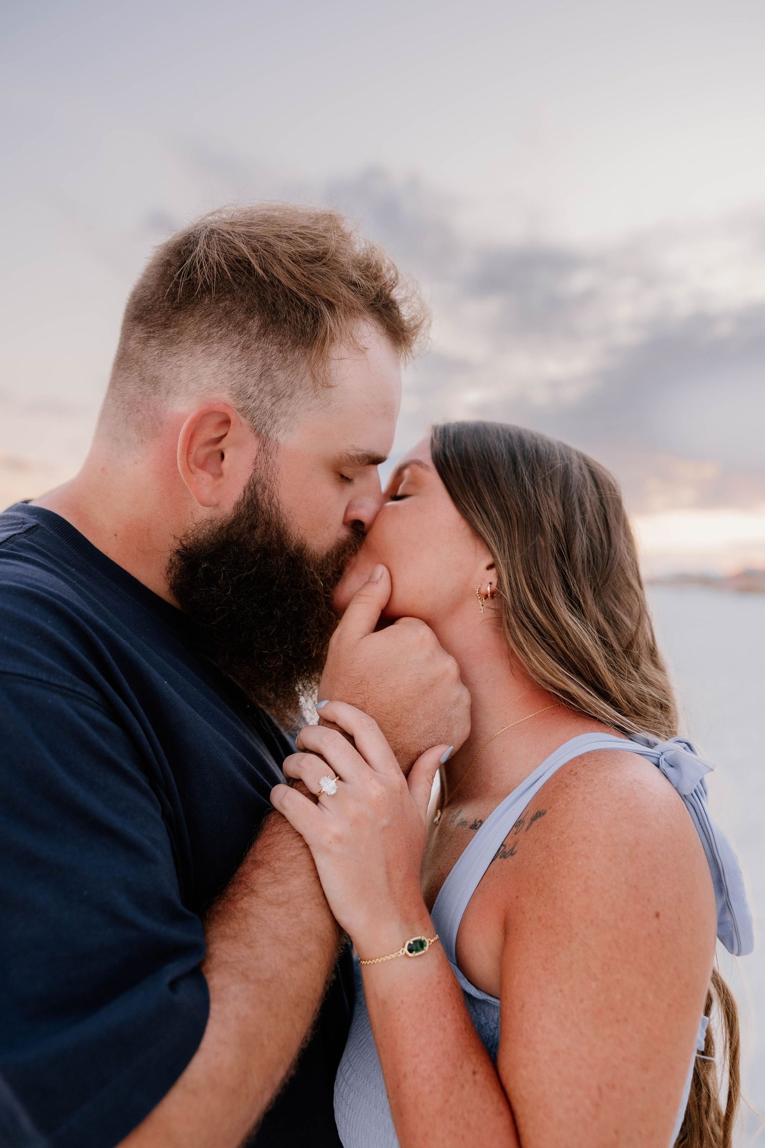 Couple sharing a romantic kiss at sunset, woman wearing engagement ring, man with beard, outdoor coastal backdrop.