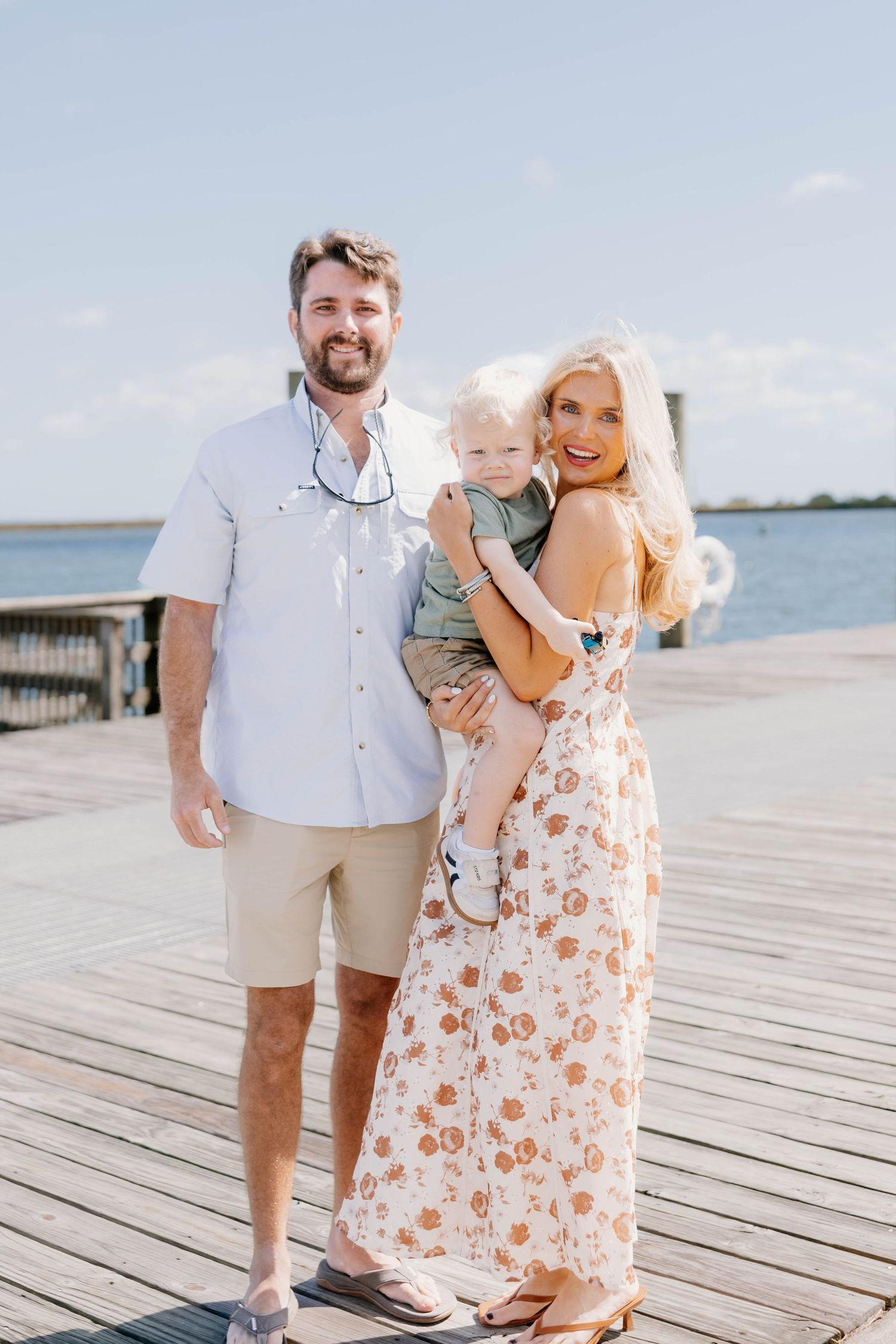 Family portrait on a sunny dock, man in white shirt and khaki shorts with blonde woman in floral maxi dress holding toddler.