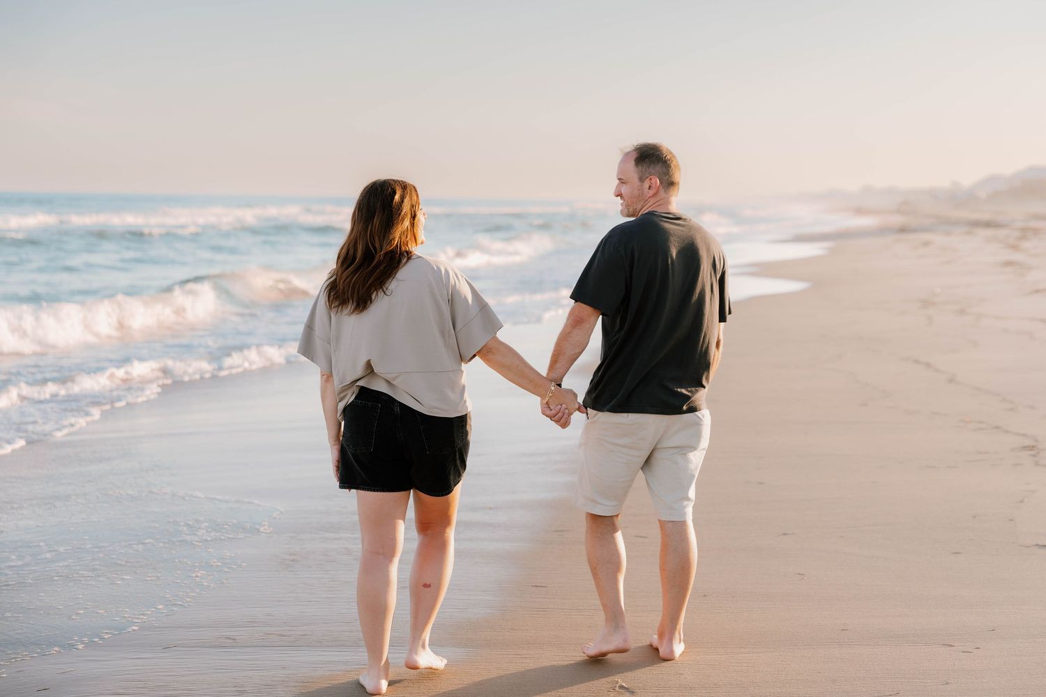 Couple holding hands walking barefoot on a sandy beach at sunset with gentle ocean waves.