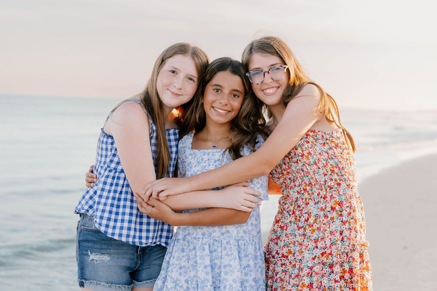 Three friends embrace while smiling on beach at sunset in summer dresses and casual wear.