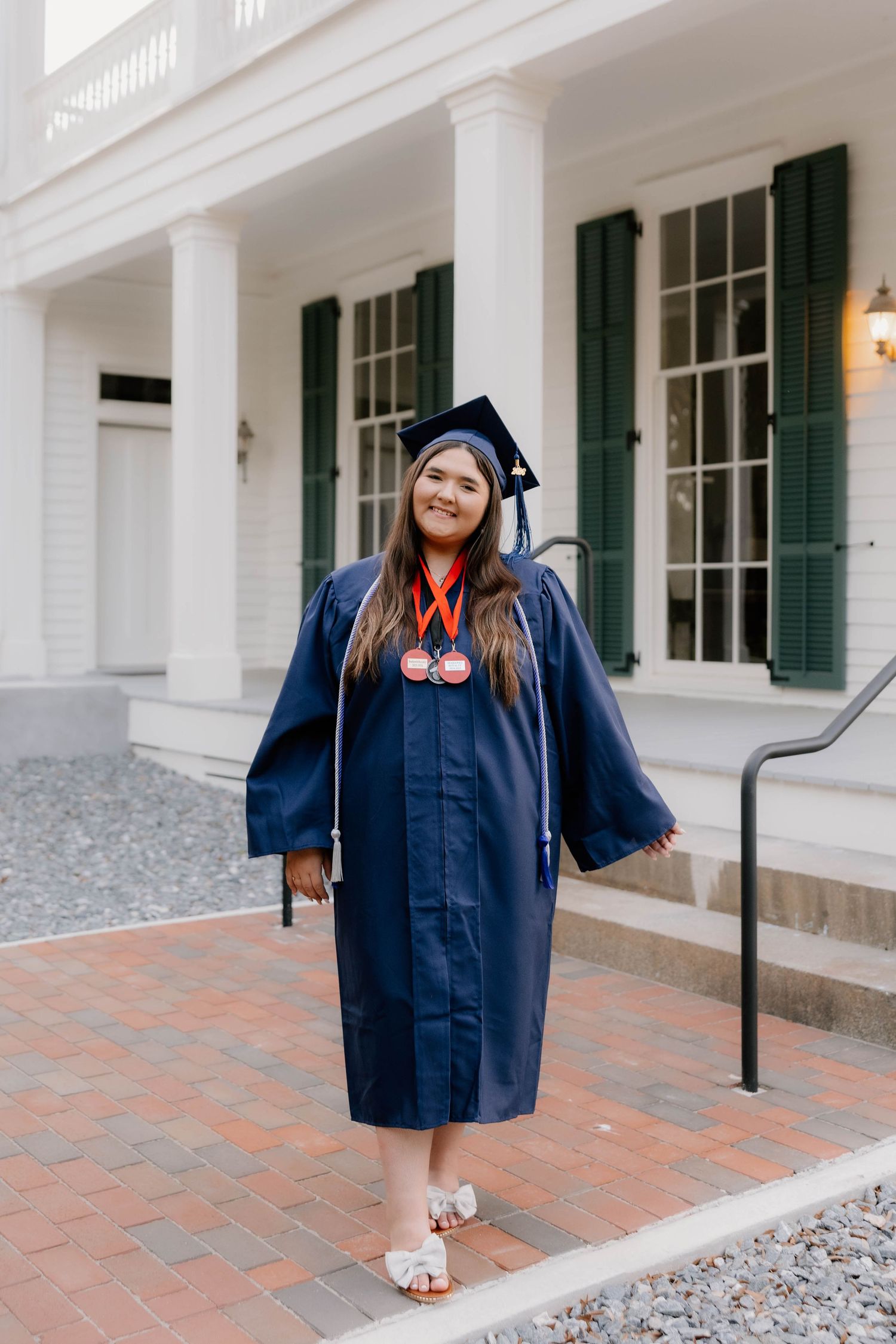 Smiling graduate in navy cap and gown with medals poses on brick path before a white colonial building.