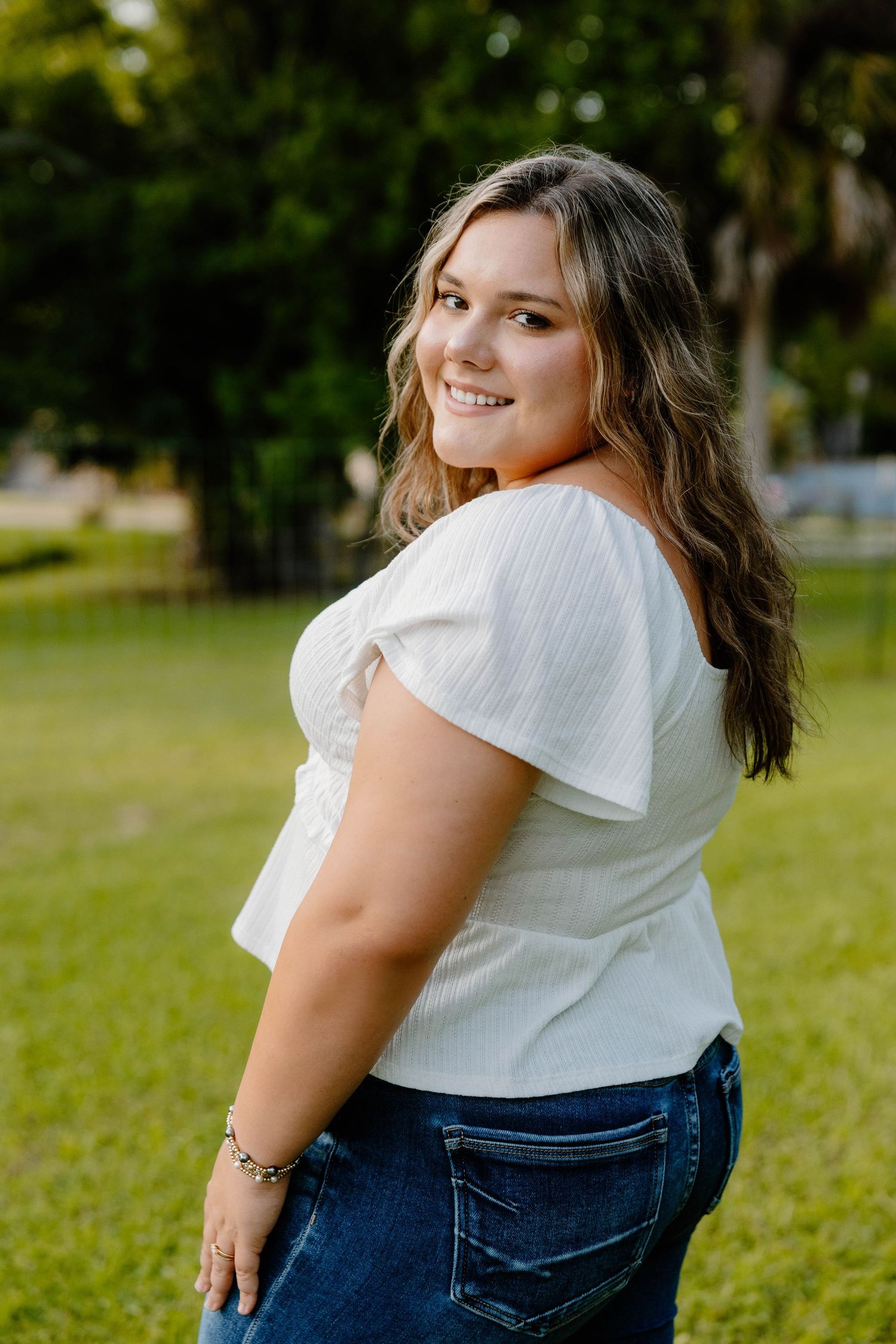 Smiling young woman in white top and jeans posing outdoors in green park setting.