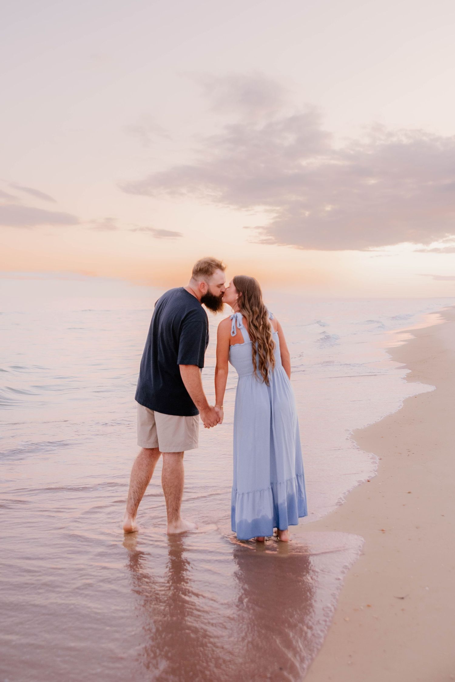 Couple kissing on a beach at sunset, man in navy shirt and shorts, woman in flowing blue dress, standing in shallow water.