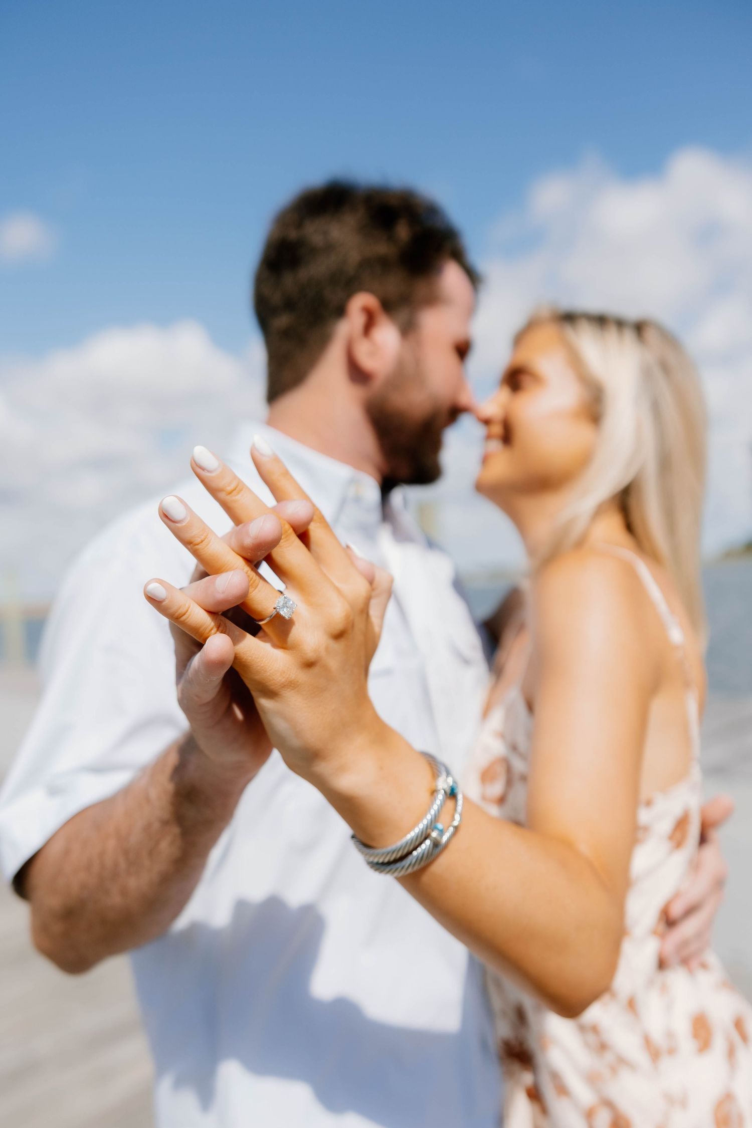 Couple sharing a romantic kiss on the beach while showing off a diamond engagement ring on a sunny day.