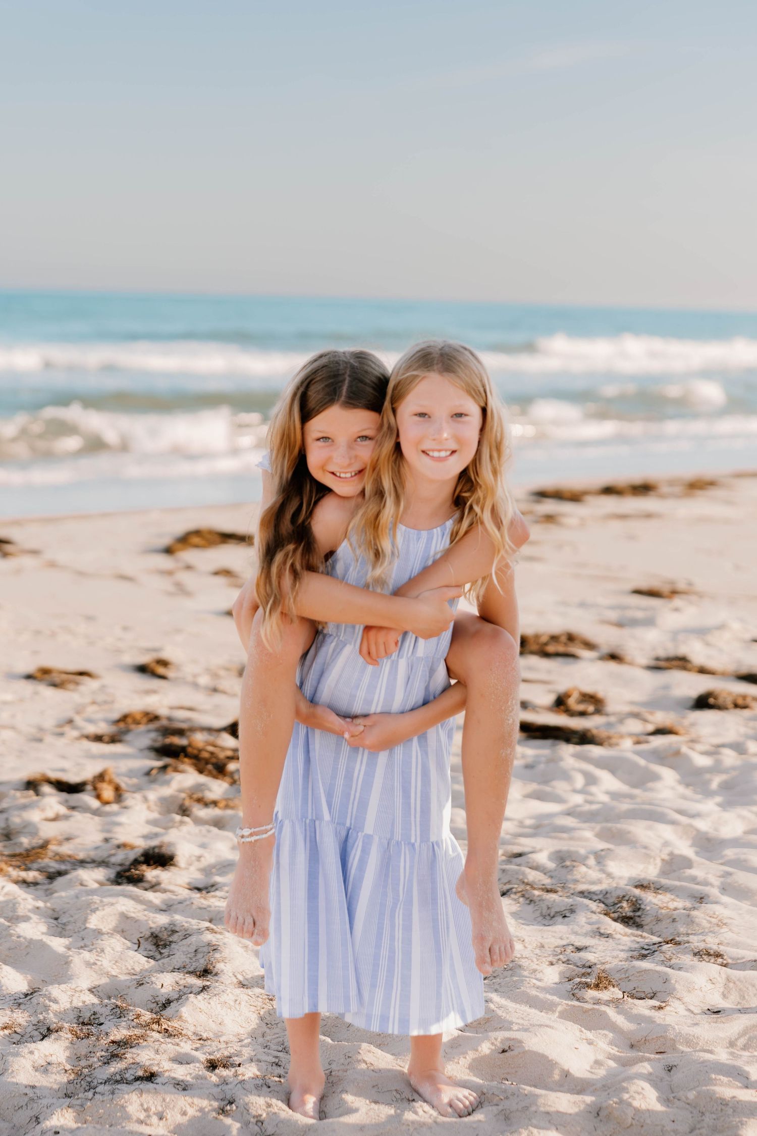 Two young girls in matching blue dresses hugging on a sandy beach with ocean waves in the background.