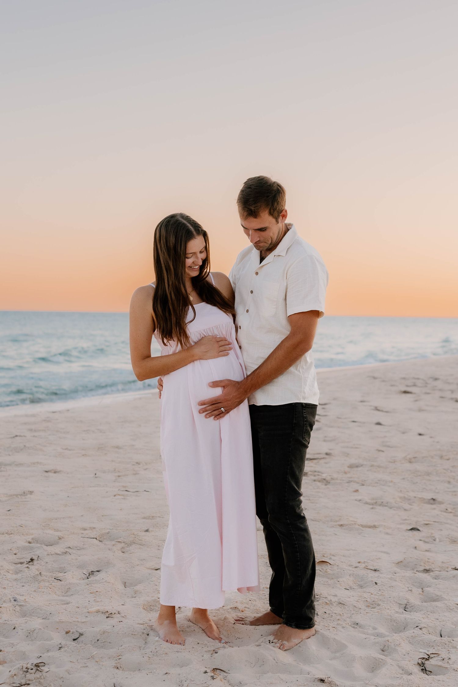 A couple stands barefoot on a beach at sunset, sharing an intimate moment while expecting a baby.