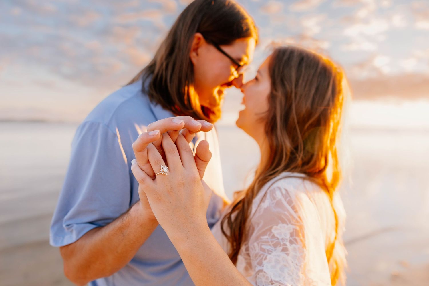 Couple sharing a romantic almost-kiss at sunset on the beach, holding hands with golden light glowing behind them.