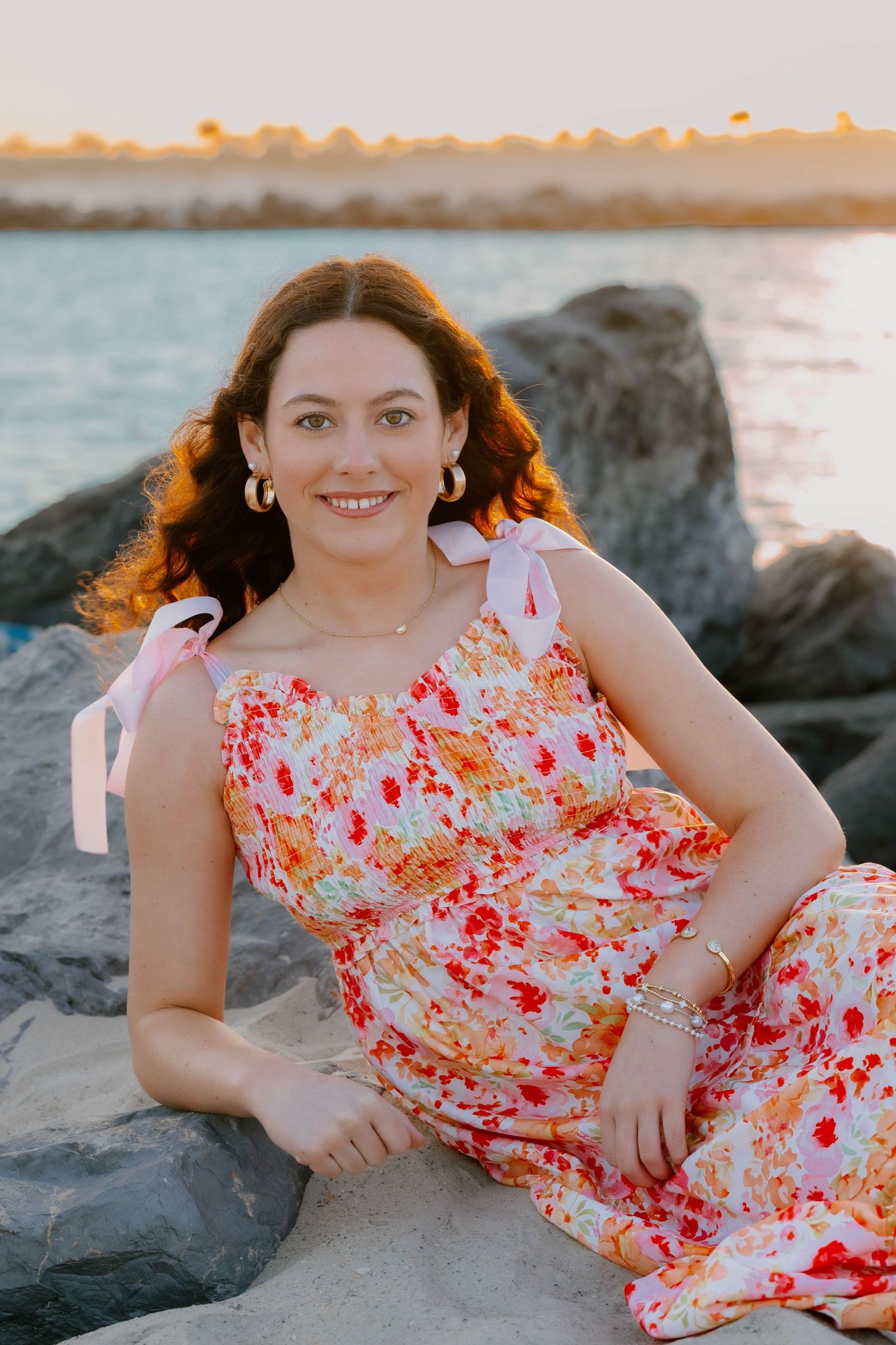 Woman in floral jumpsuit sitting on rocks by the ocean at sunset, smiling warmly, wearing gold earrings and pink bow straps.