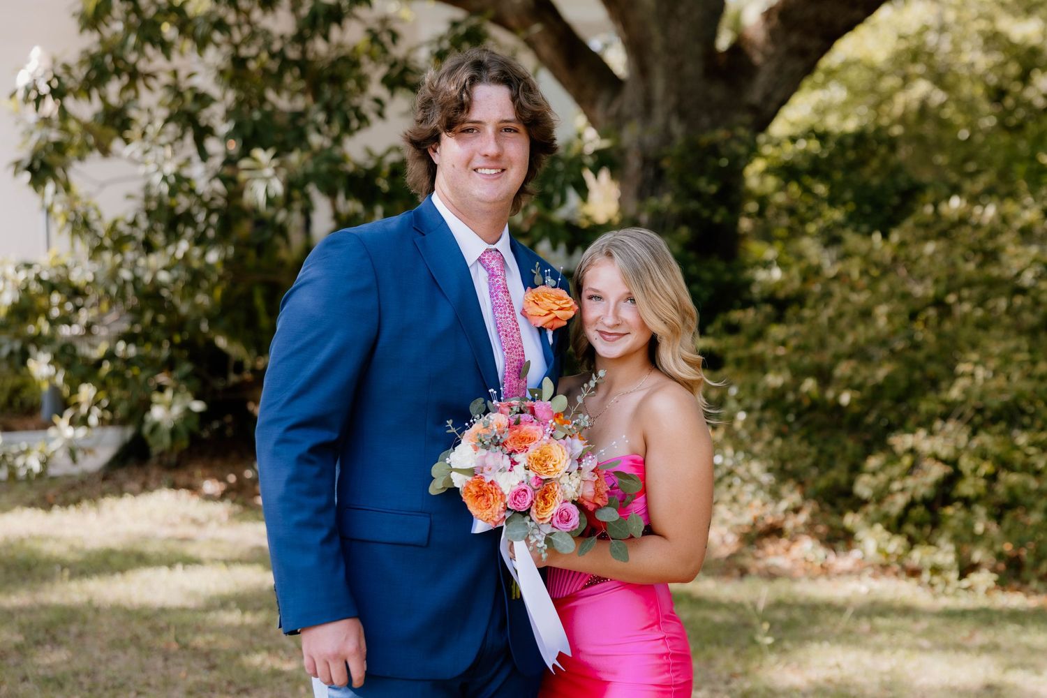 A young couple dressed in formal attire poses outdoors, the girl in a pink dress holding a colorful bouquet.