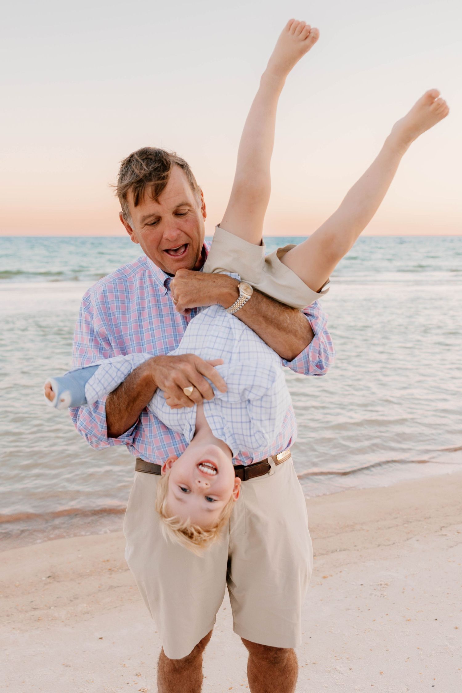 Man holding laughing child upside down on a sandy beach at sunset, both smiling and enjoying playful family moment.