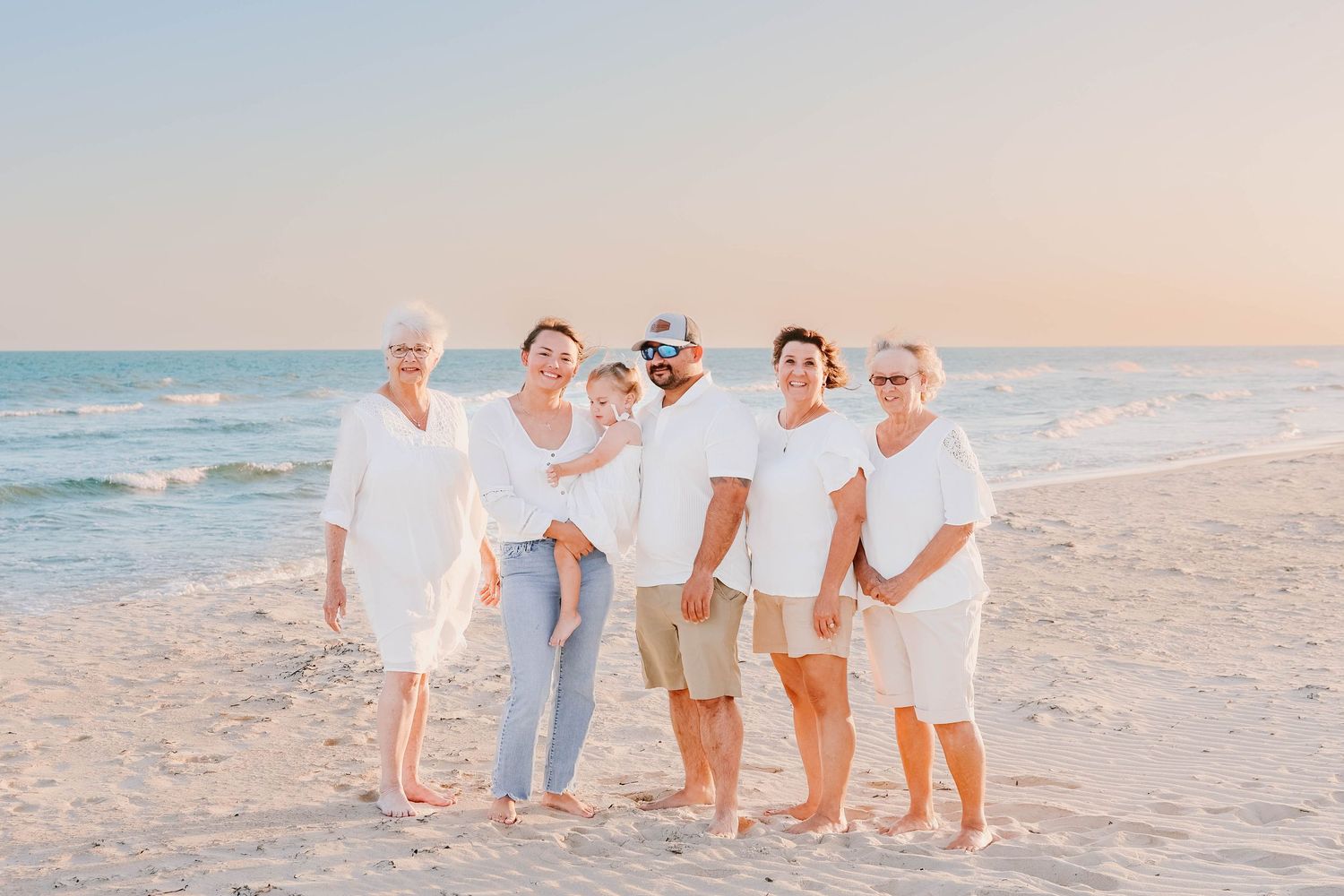 A family wearing coordinated white and beige outfits walks together on a sandy beach at sunset.