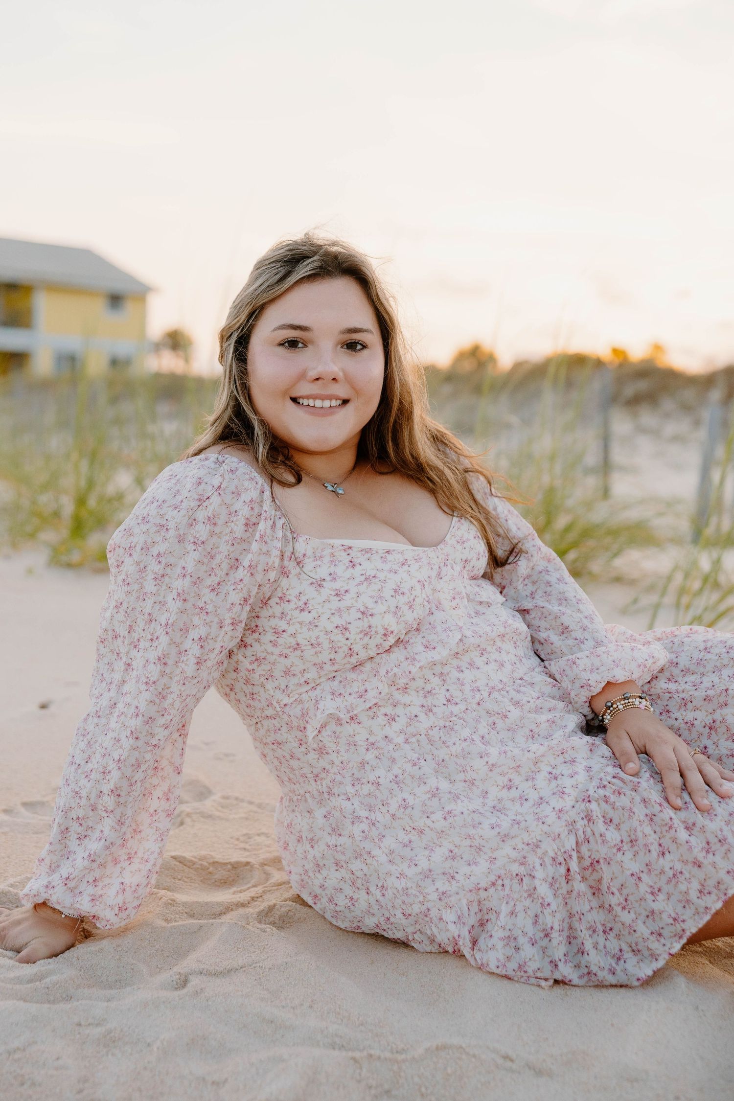Young woman in floral dress sitting on sandy beach at sunset smiling at camera.