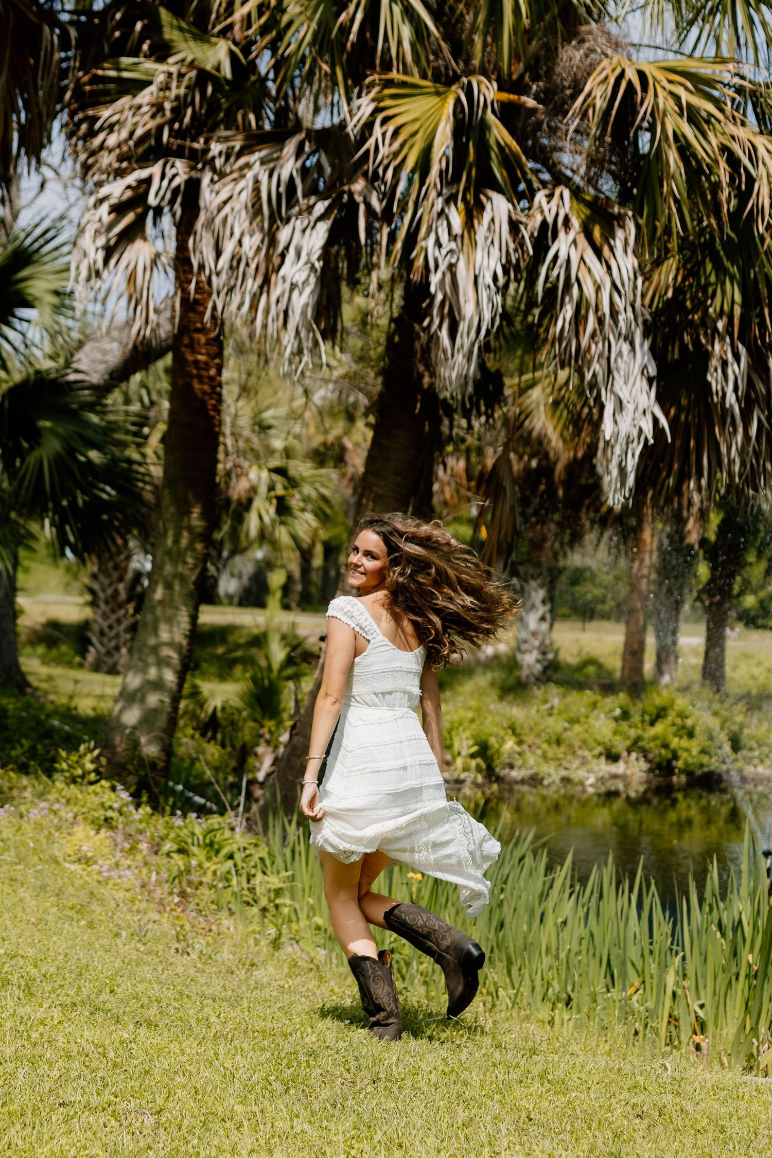 Woman in white dress and cowboy boots twirling on green grass with palm trees and pond in background.
