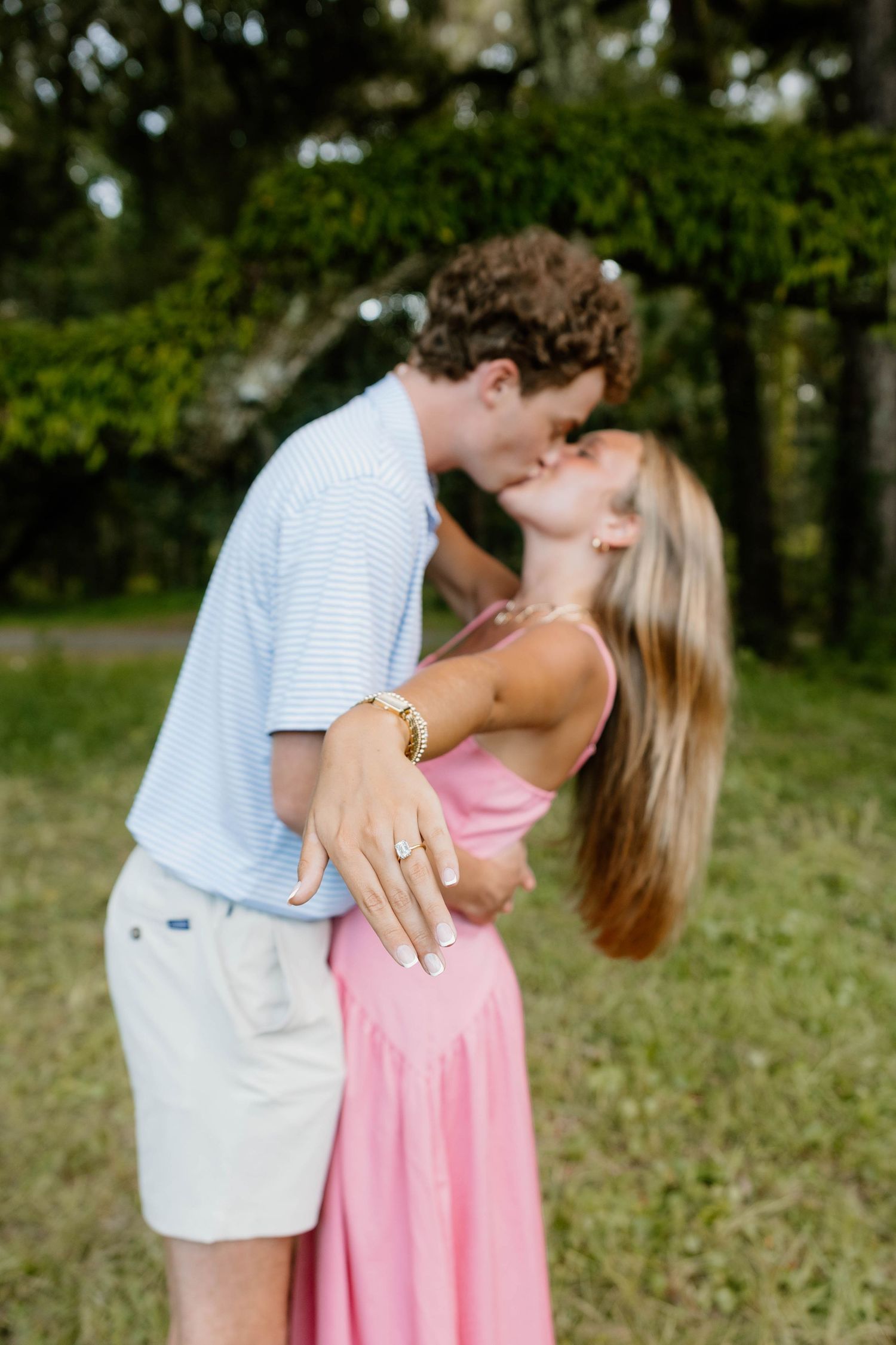 Couple sharing a romantic kiss outdoors, woman displaying engagement ring in pink dress, lush green trees in background.