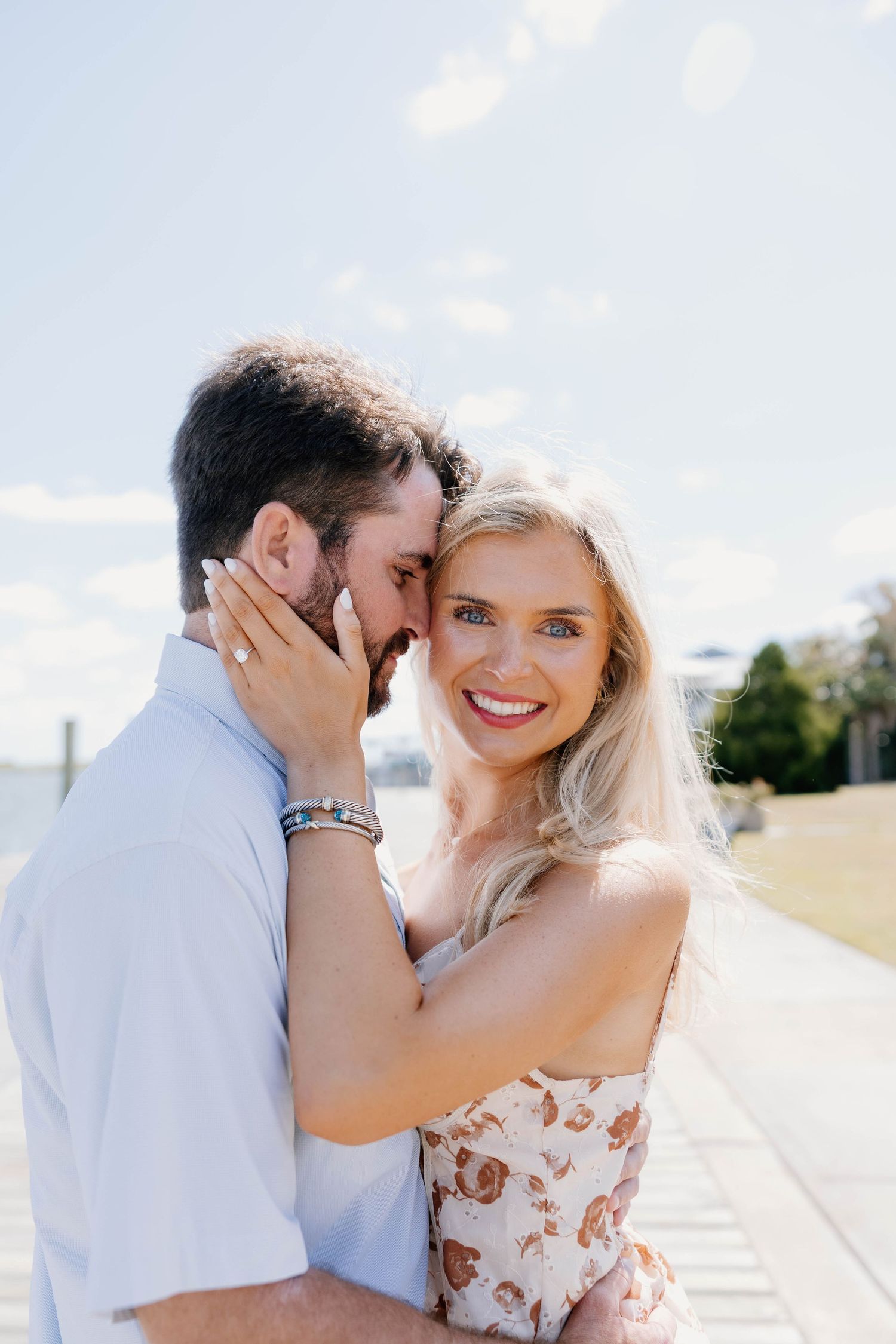 Smiling blonde woman in floral dress embraces dark-haired bearded man on sunny waterfront boardwalk during engagement photo session.