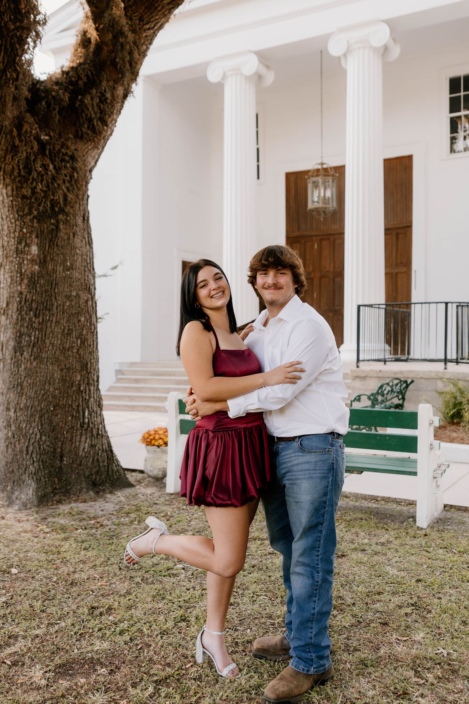 A smiling couple embraces in front of a white colonial building, woman in burgundy dress, man in white shirt and jeans.