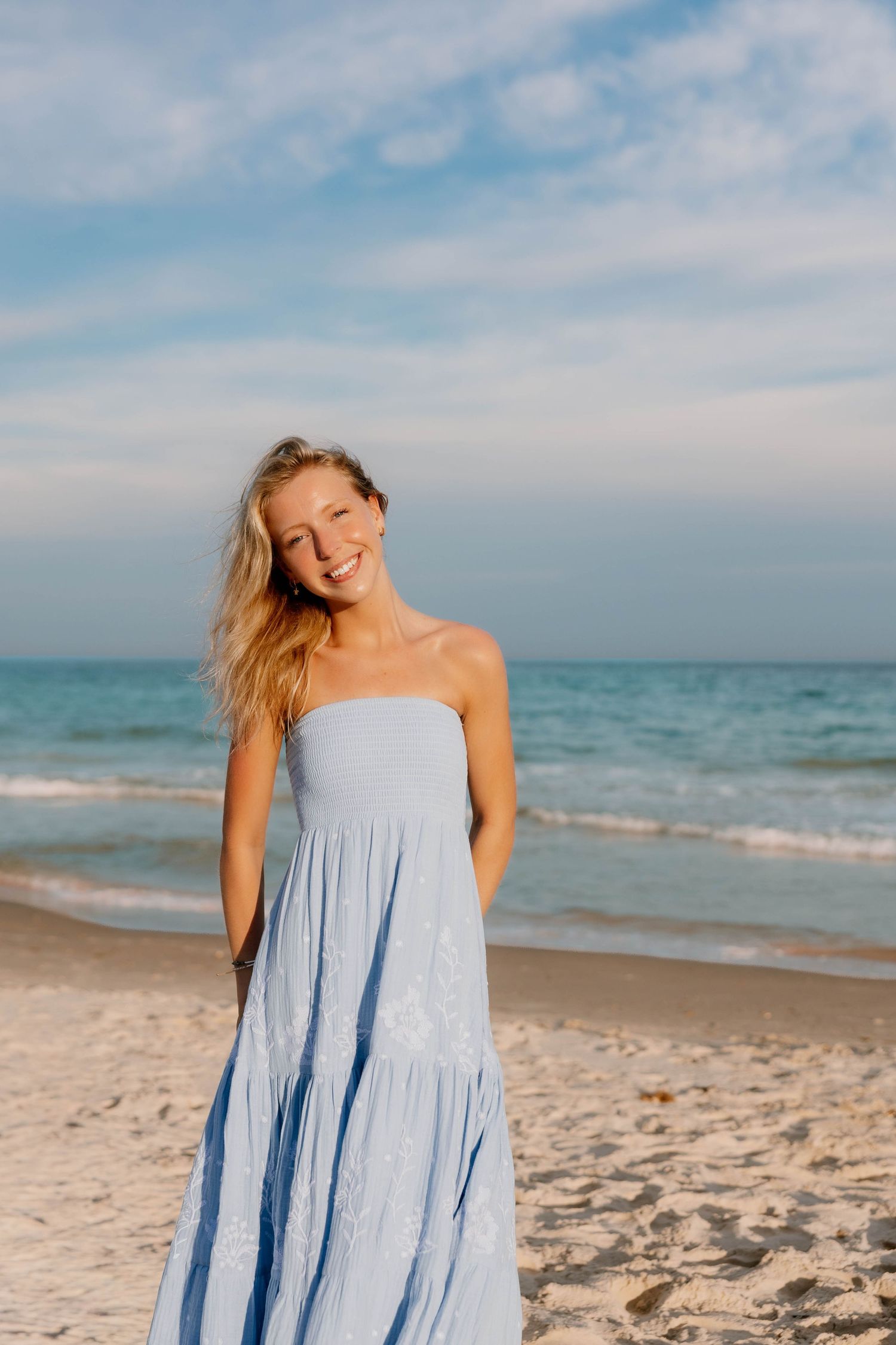 Smiling blonde woman in a light blue strapless maxi dress standing on a sandy beach with ocean waves behind her.