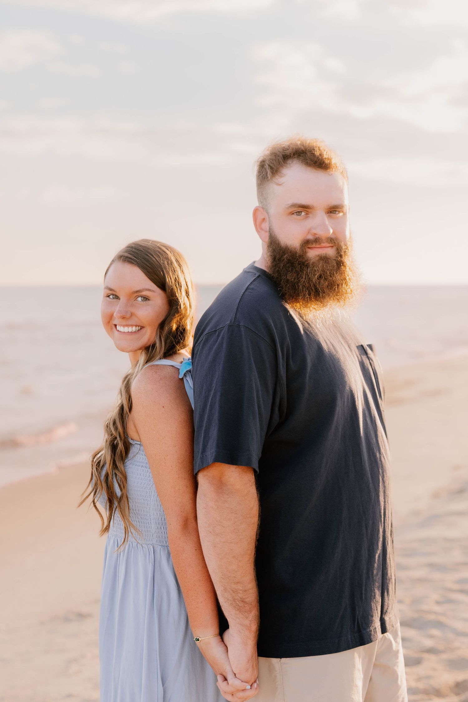 A couple stands together on a sandy beach during sunset, wearing blue dress and black shirt.
