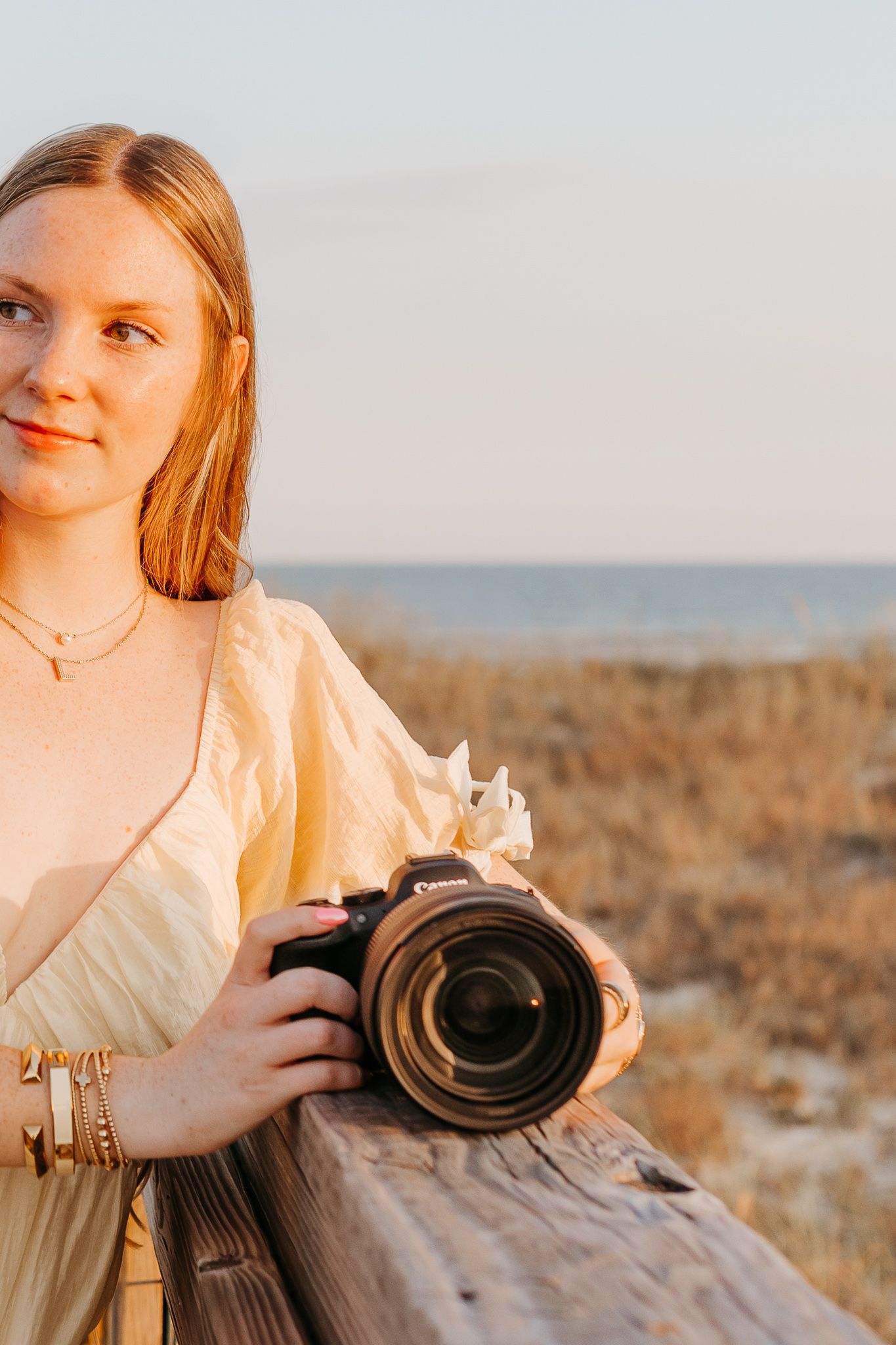 Young woman holding a DSLR camera at a sunny beach, wearing a yellow dress and gold jewelry.