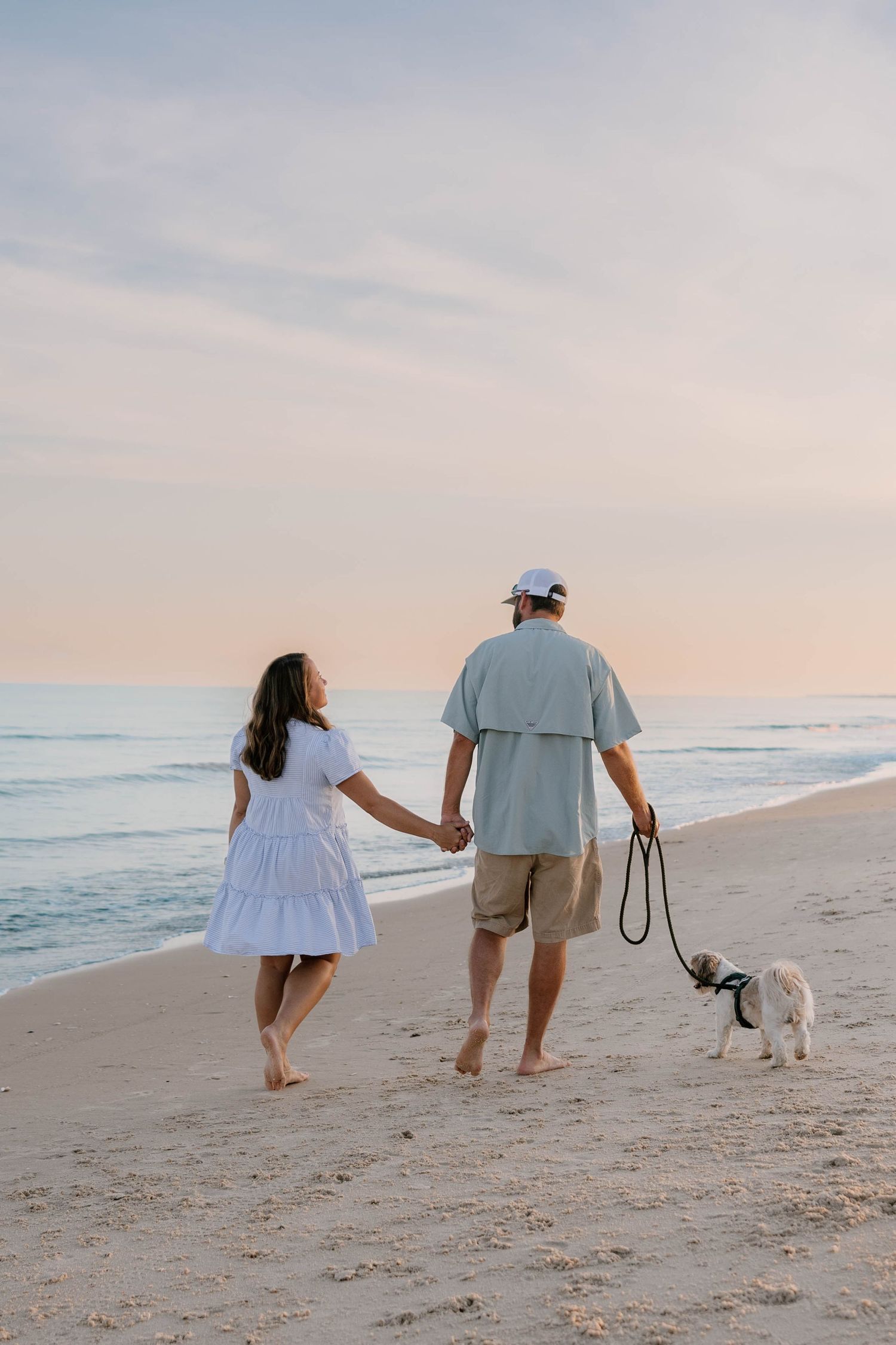 Couple walks dog on beach at sunset while holding hands.
