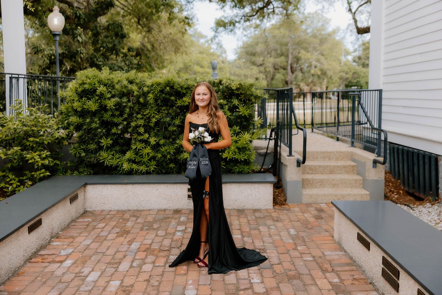 Young woman in elegant black gown holding bouquet poses on brick courtyard with lush greenery and white building steps.