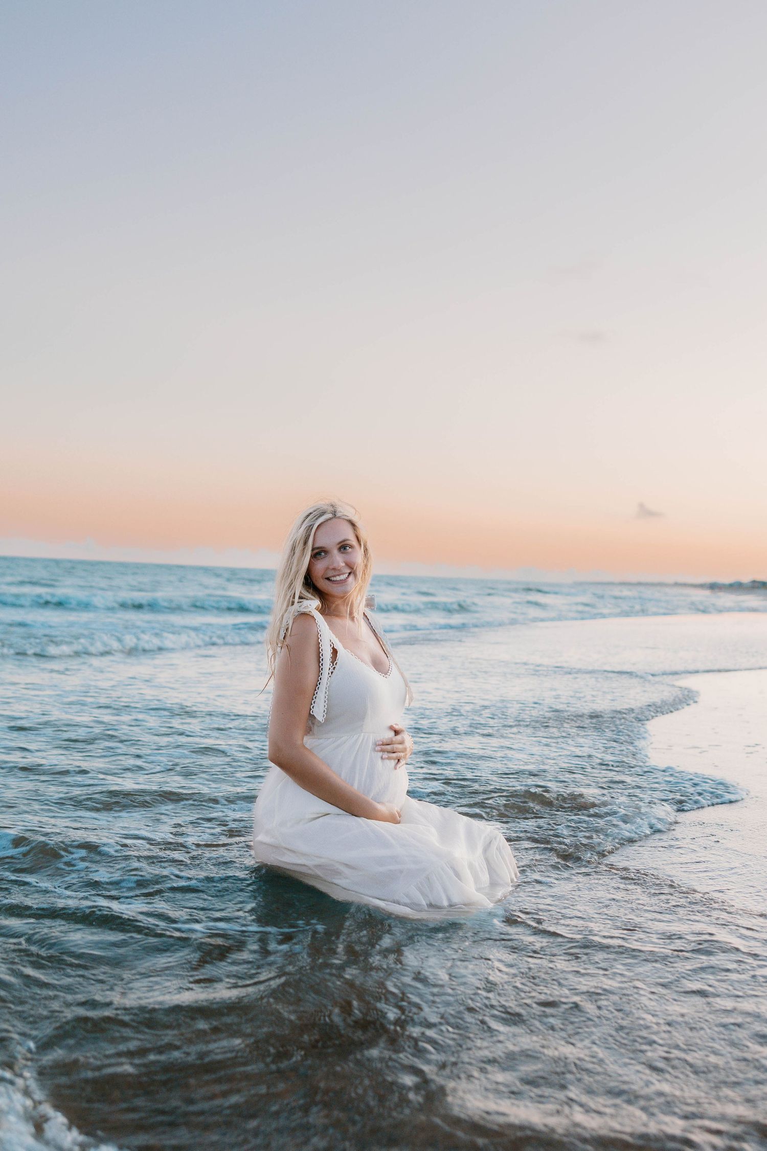 Smiling pregnant woman in white dress sitting in ocean waves at sunset beach.