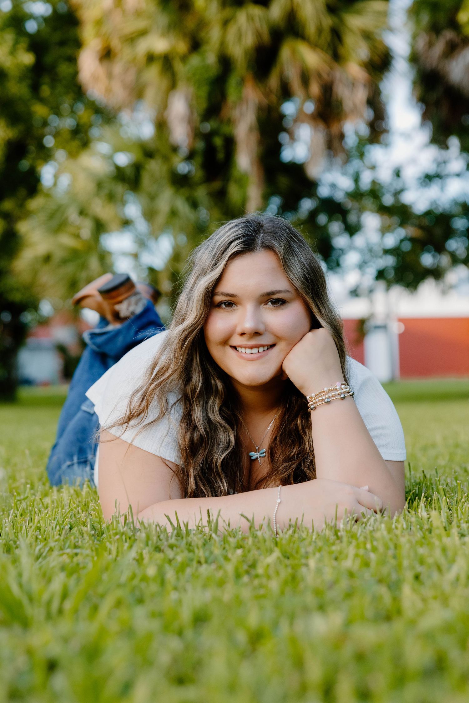 Young woman with wavy hair lying on green grass, smiling, wearing a white top and jeans outdoors.