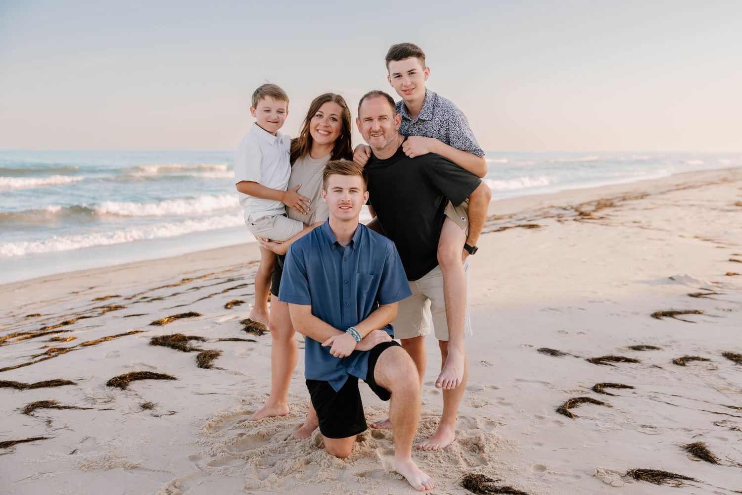 Family of five smiling together on a sandy beach at sunset, with ocean waves in the background.