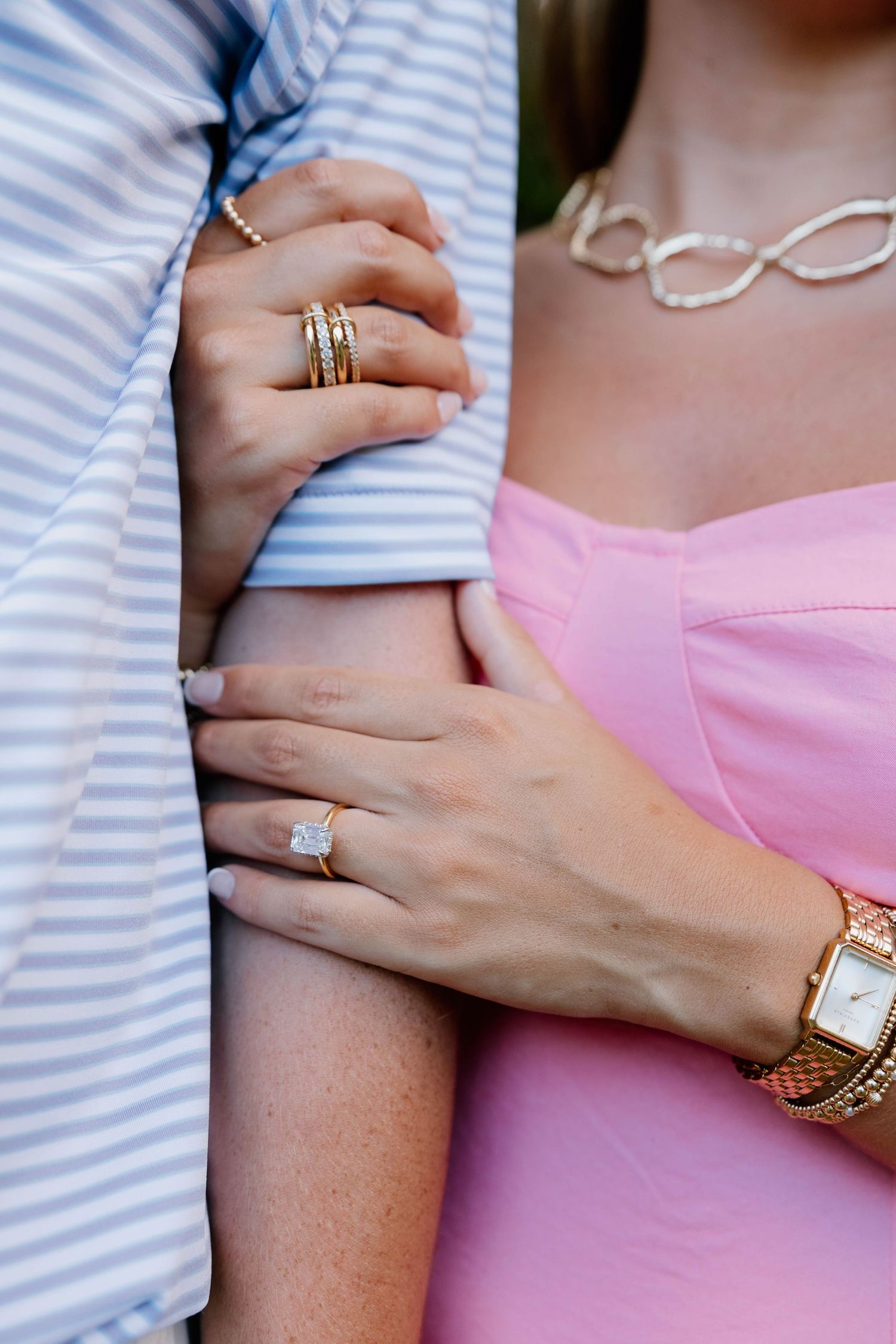 Close-up of a woman in pink dress wearing a diamond engagement ring, gold watch, and bracelet holding a man's arm.