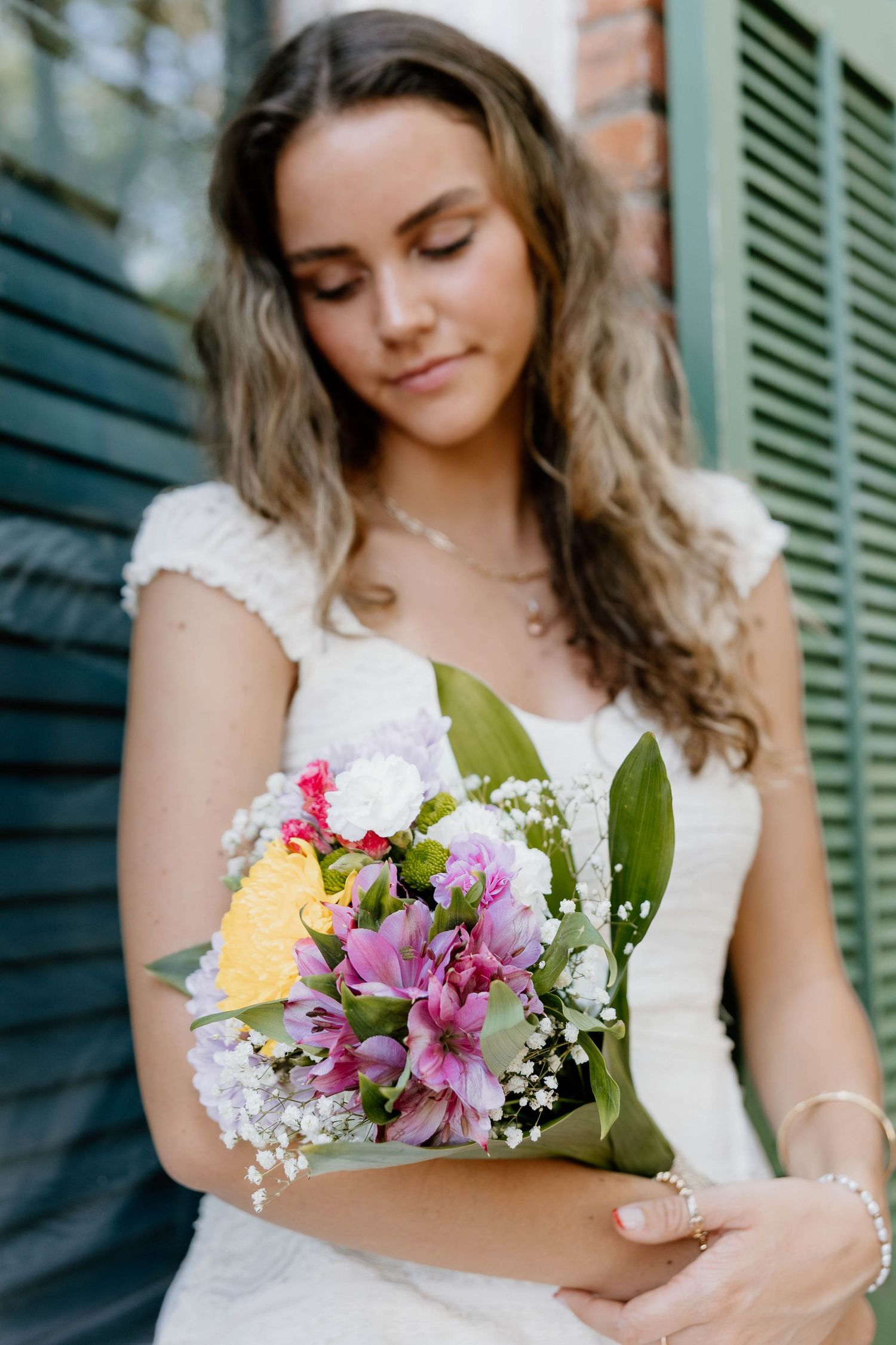 Young bride in white lace dress holding colorful bouquet with purple alstroemeria and yellow roses, looking down softly.