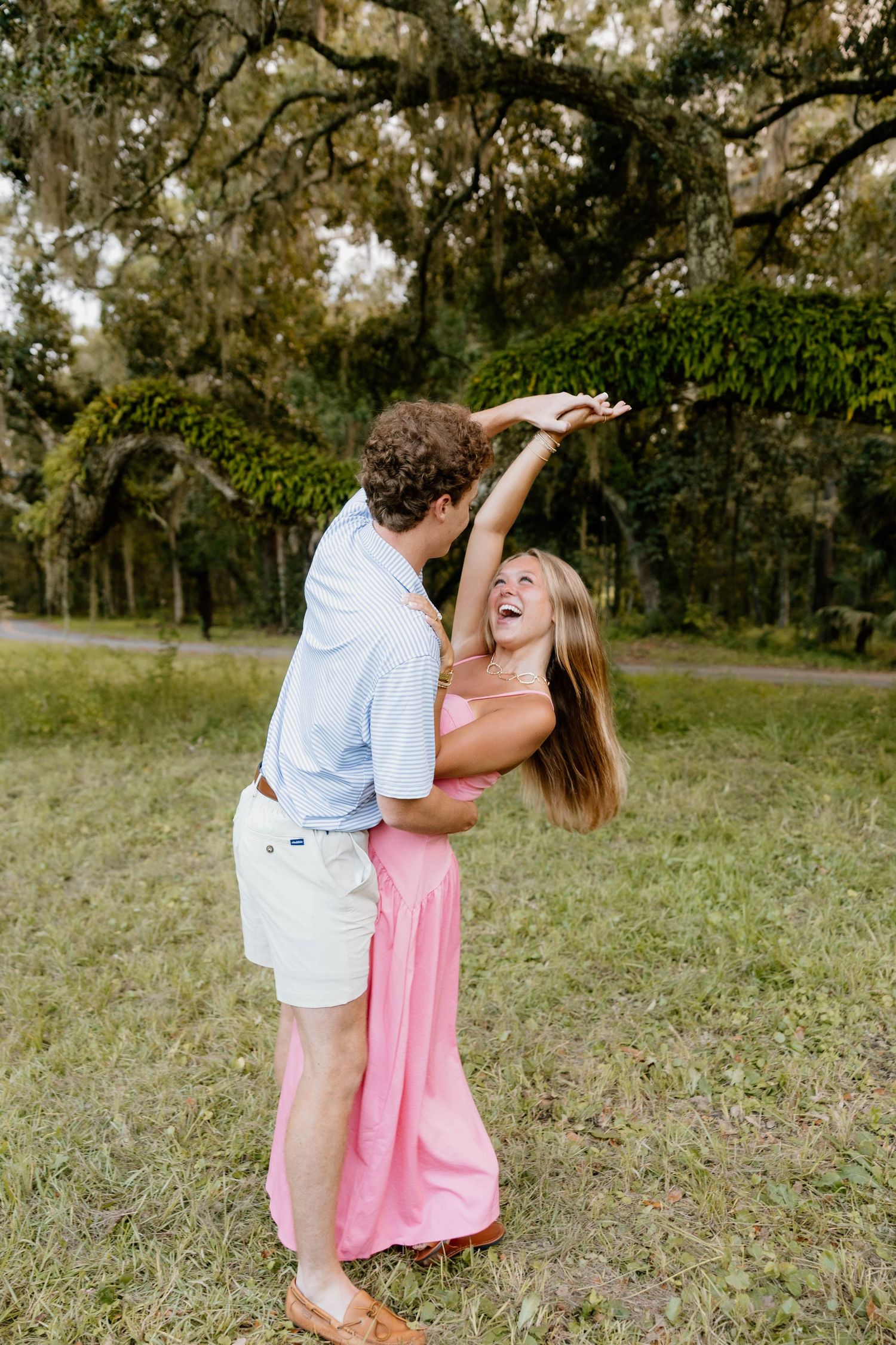 A joyful couple dancing outdoors, woman in pink dress being twirled under oak trees on a grassy lawn.