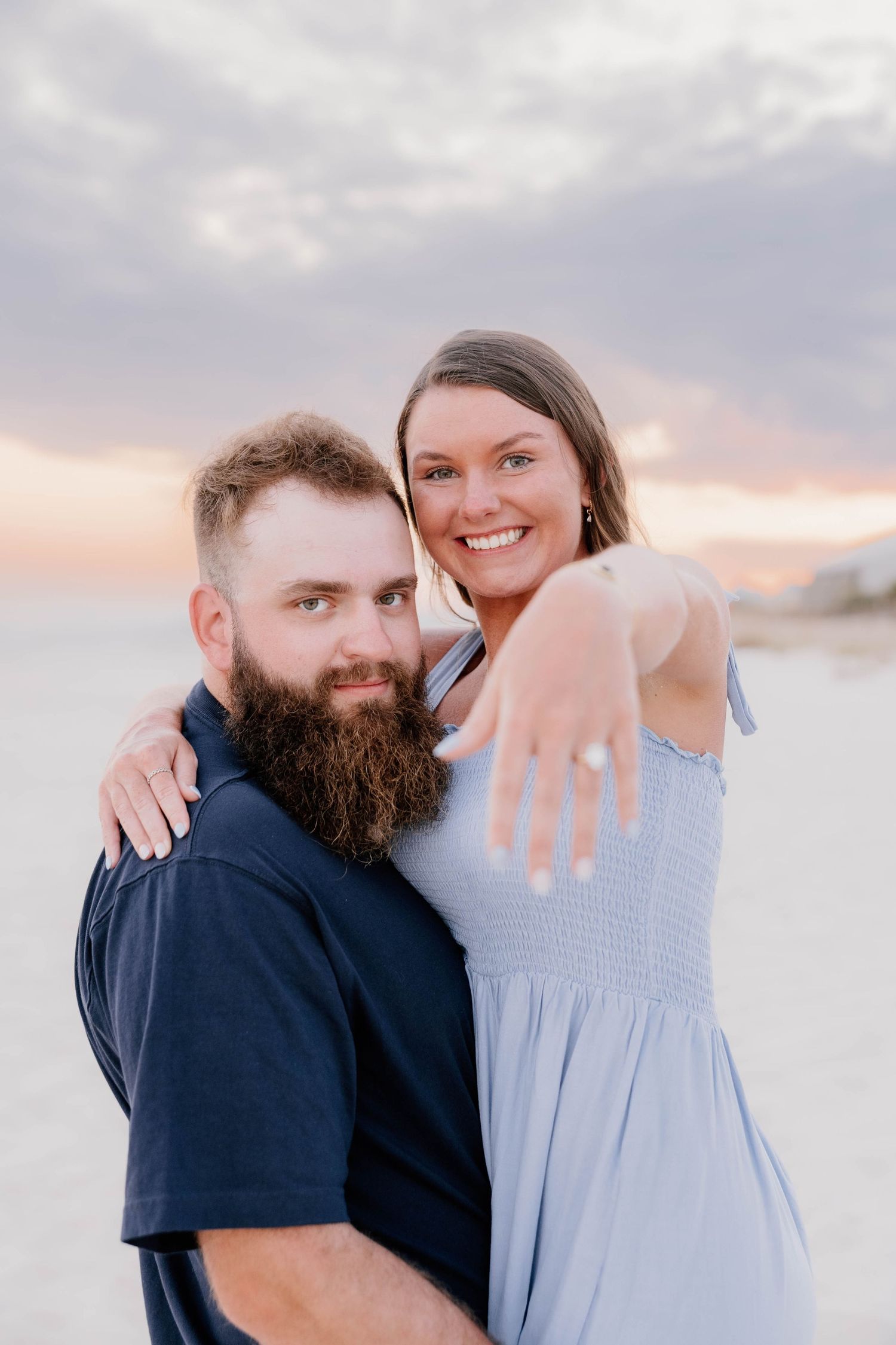 A couple embraces on a beach at sunset while showing off an engagement ring.