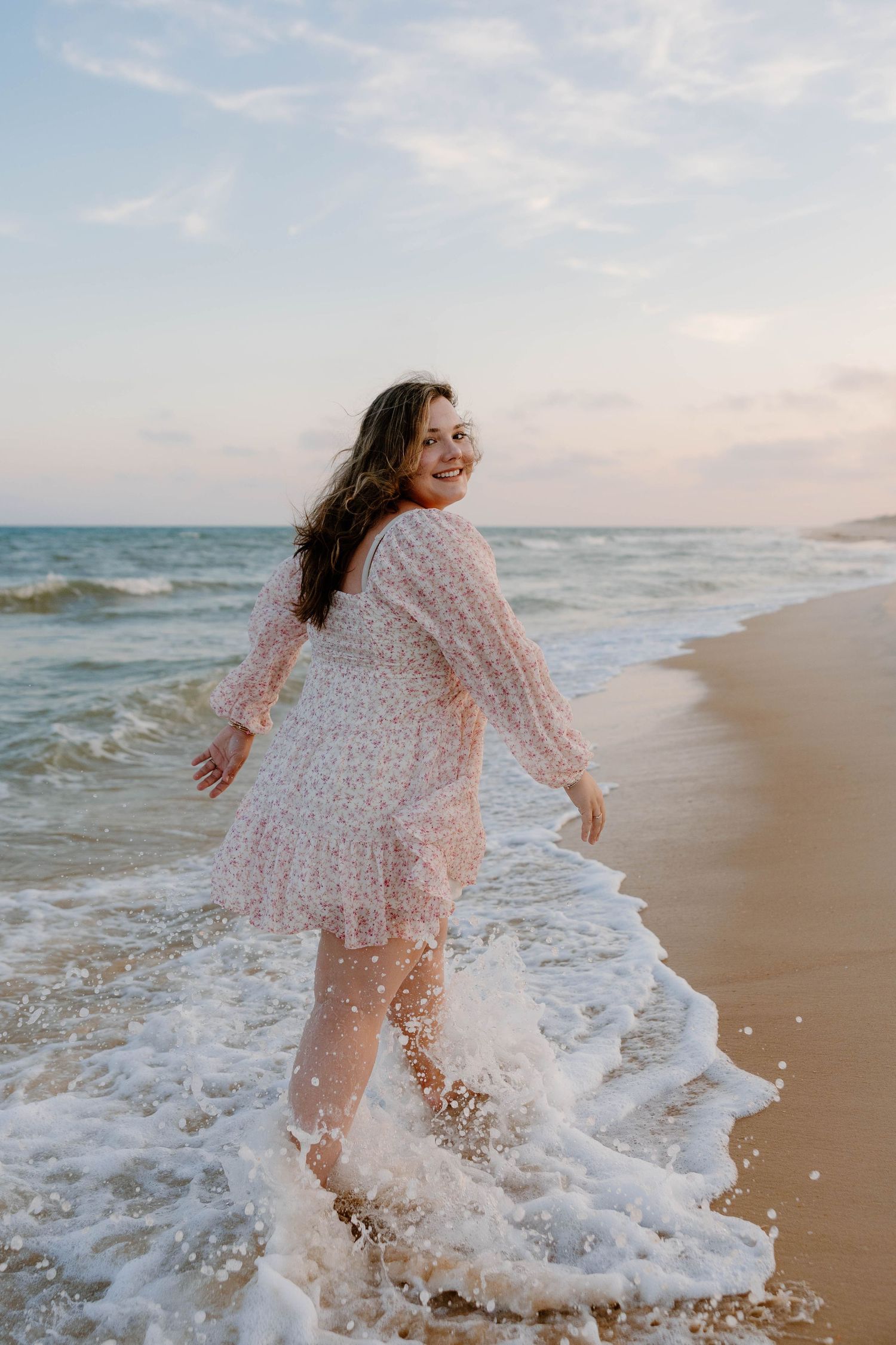 A smiling woman in a floral dress walks along a sandy beach as ocean waves splash around her feet at sunset.