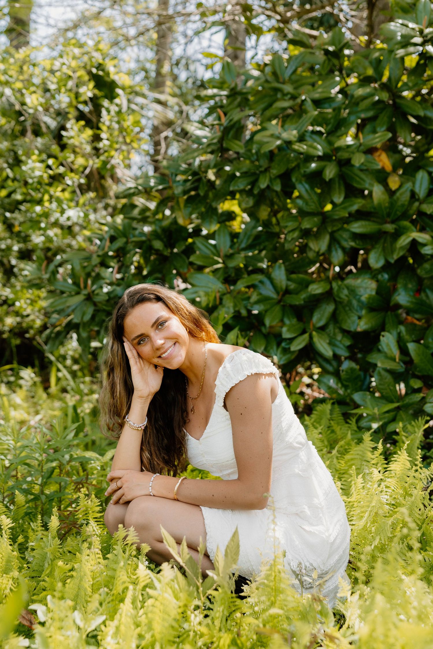 Young woman in white dress smiling while sitting among lush green ferns and shrubs in a garden setting.