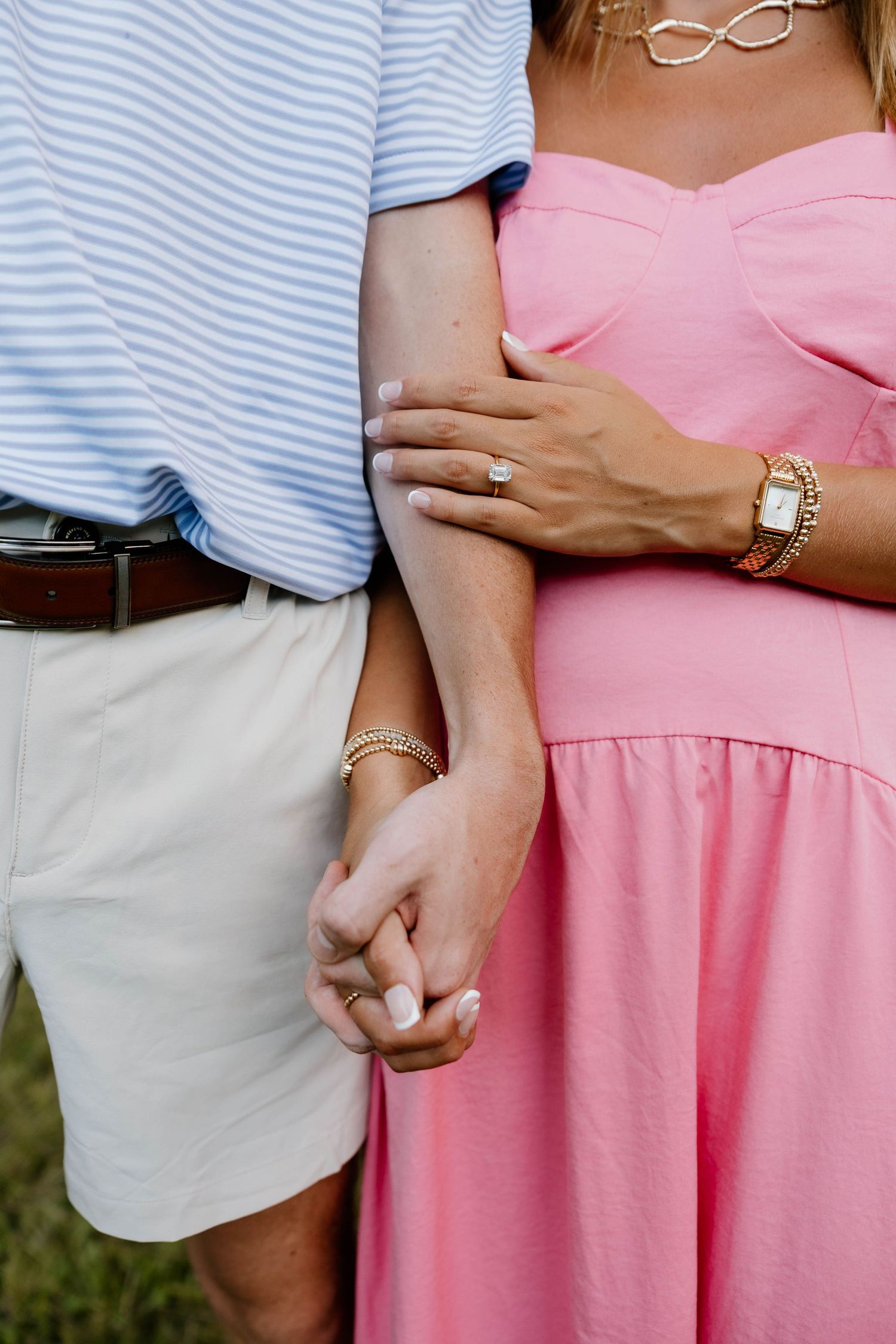 Couple holding hands, woman in pink dress with engagement ring and gold bracelet, man in blue striped shirt and khaki shorts.