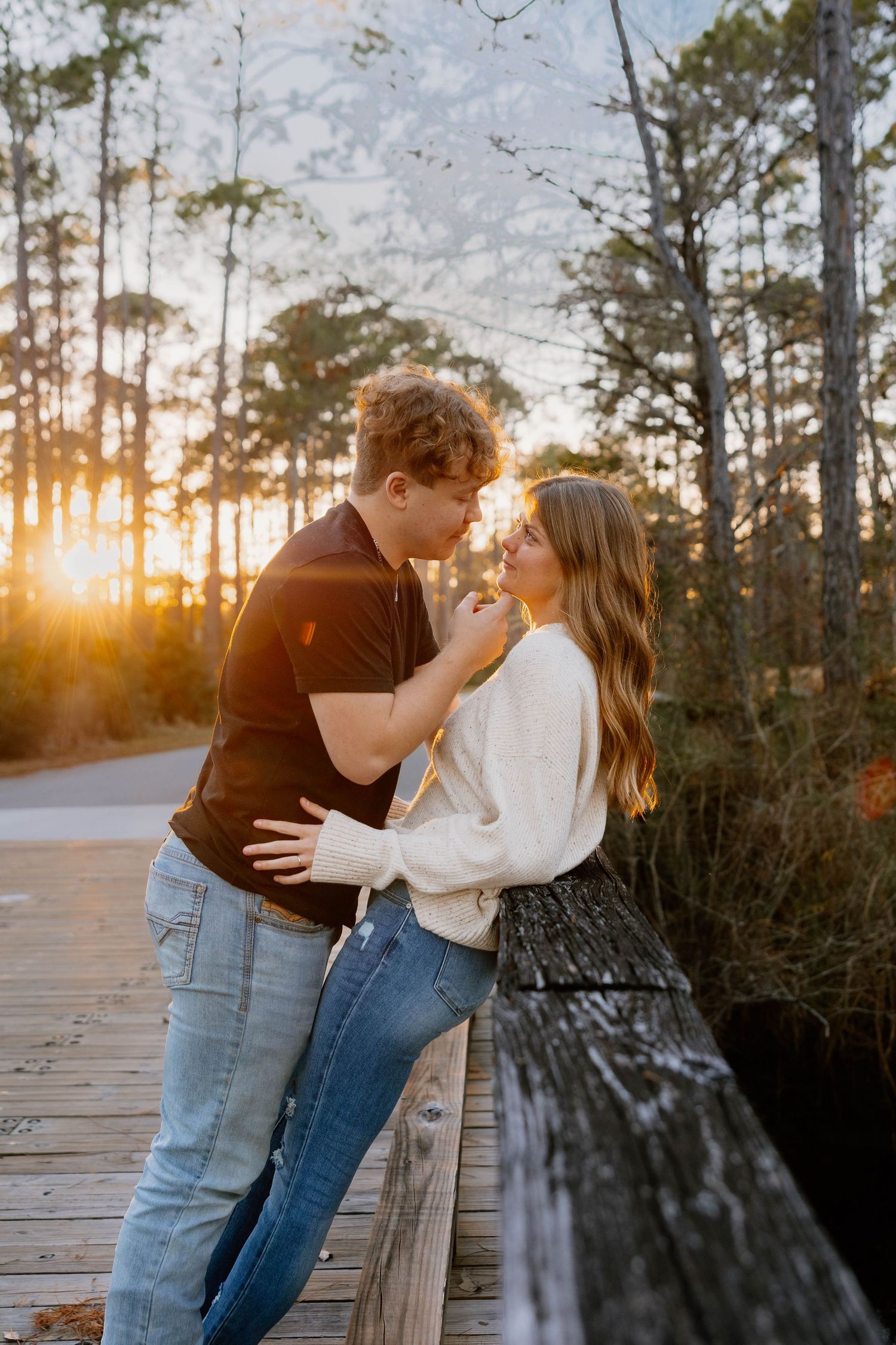 Couple sharing a romantic moment on a wooden boardwalk at golden sunset, surrounded by tall pine trees.
