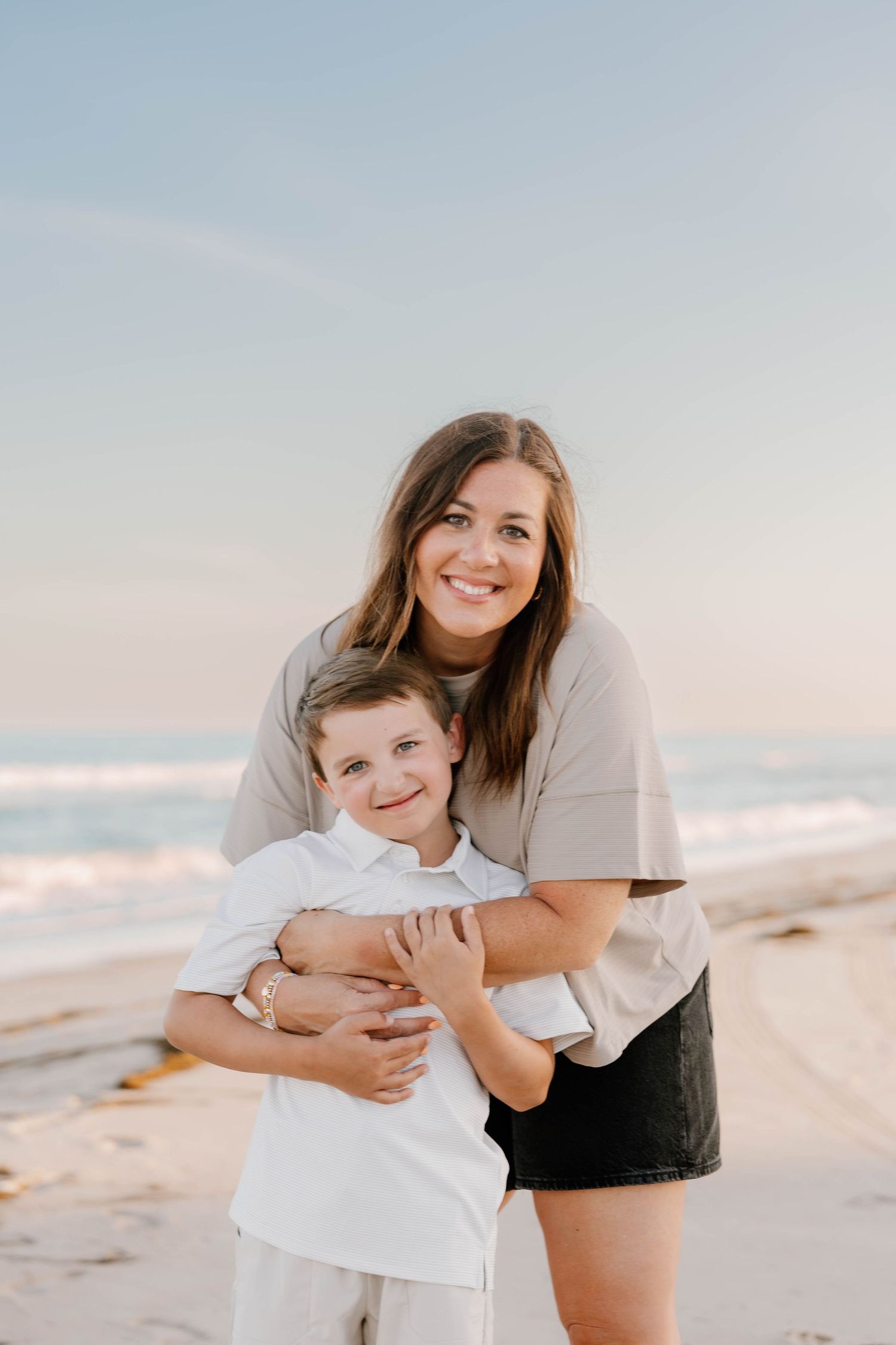 A woman hugging a young boy on a sunny beach, both smiling at the camera with ocean waves in the background.