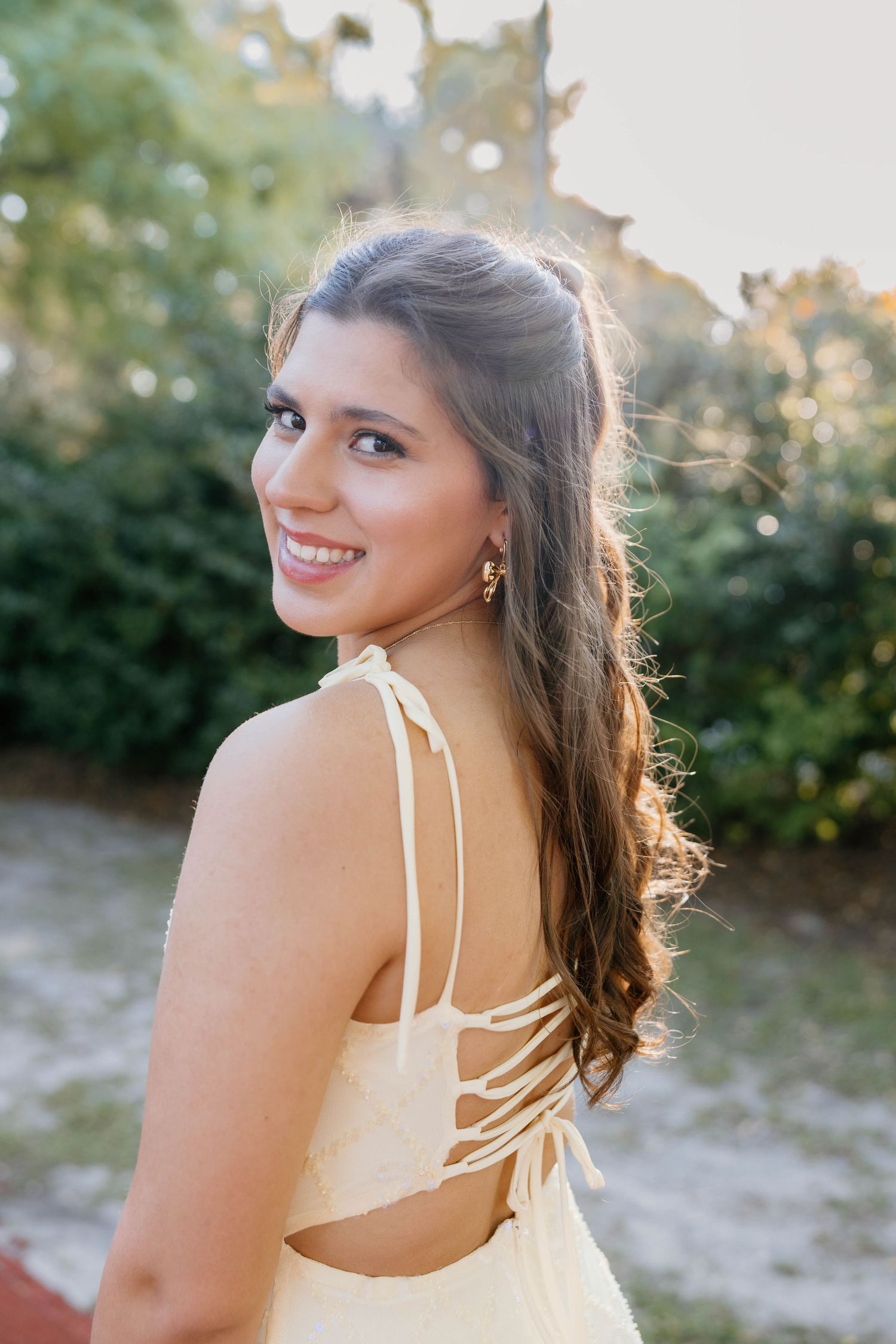 Young woman in yellow sundress smiling over shoulder outdoors in golden sunlight with lush green trees background.