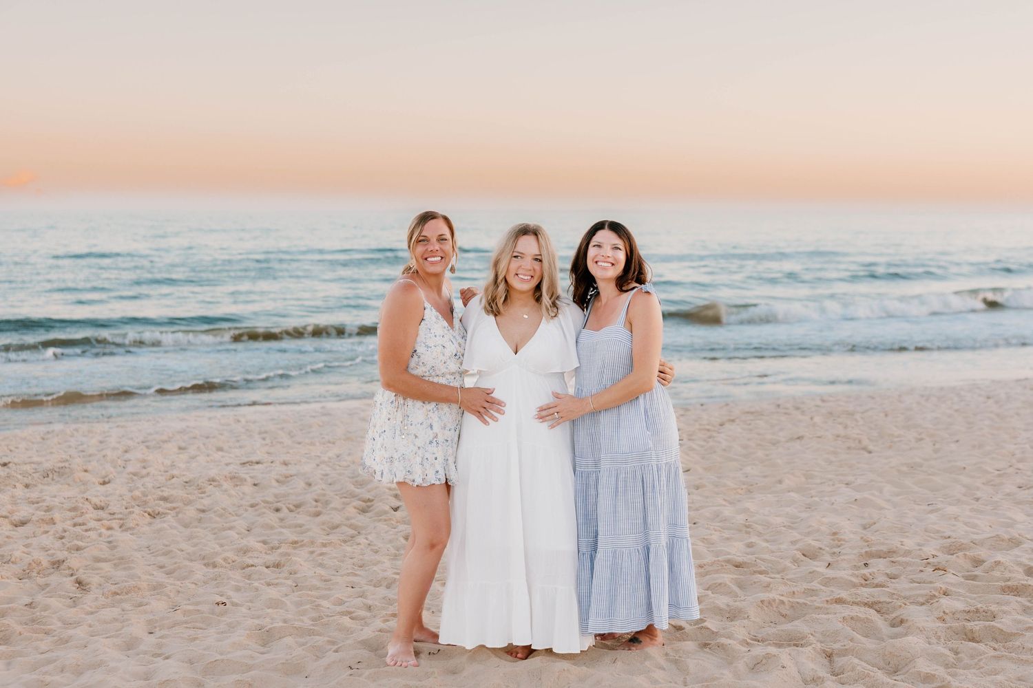 Three women pose together on a sandy beach at sunset, with the middle woman in a white dress cradling a baby bump.