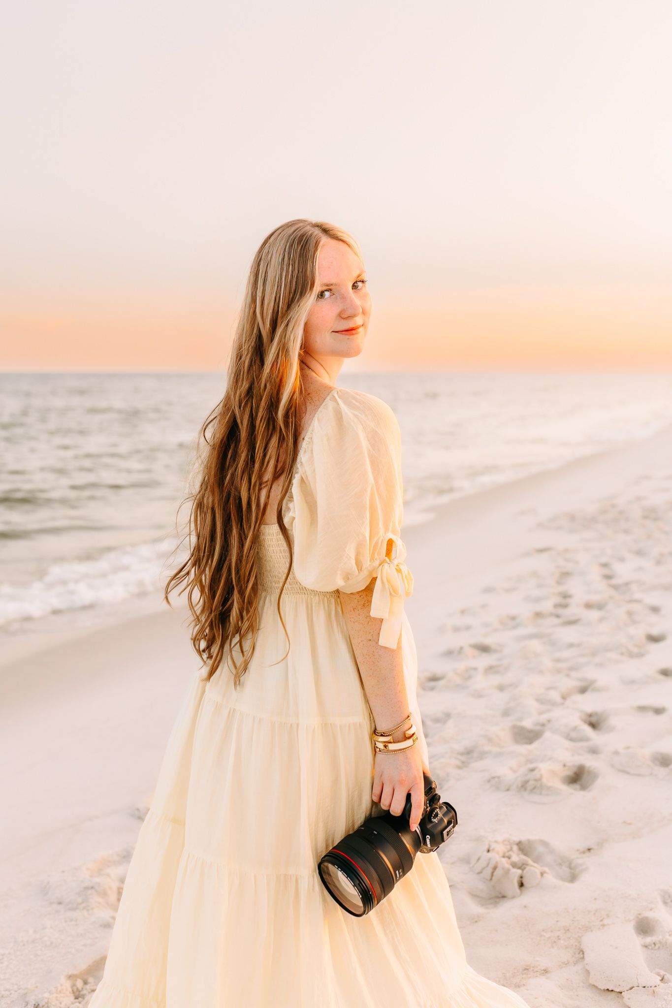 Young woman in cream dress holding camera on sandy beach at golden hour sunset.