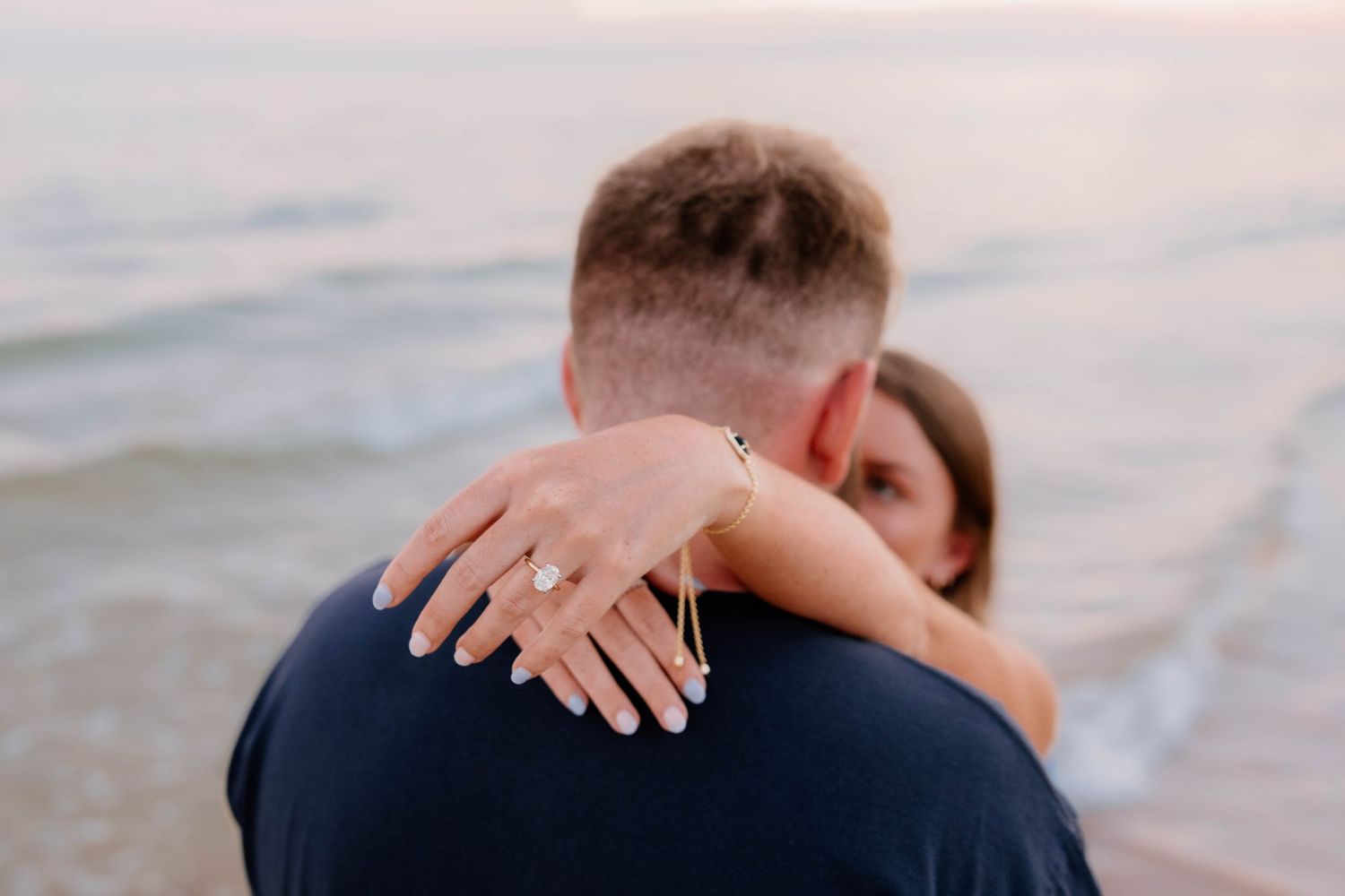 Couple embracing on beach with woman showing diamond engagement ring on her hand at sunset.