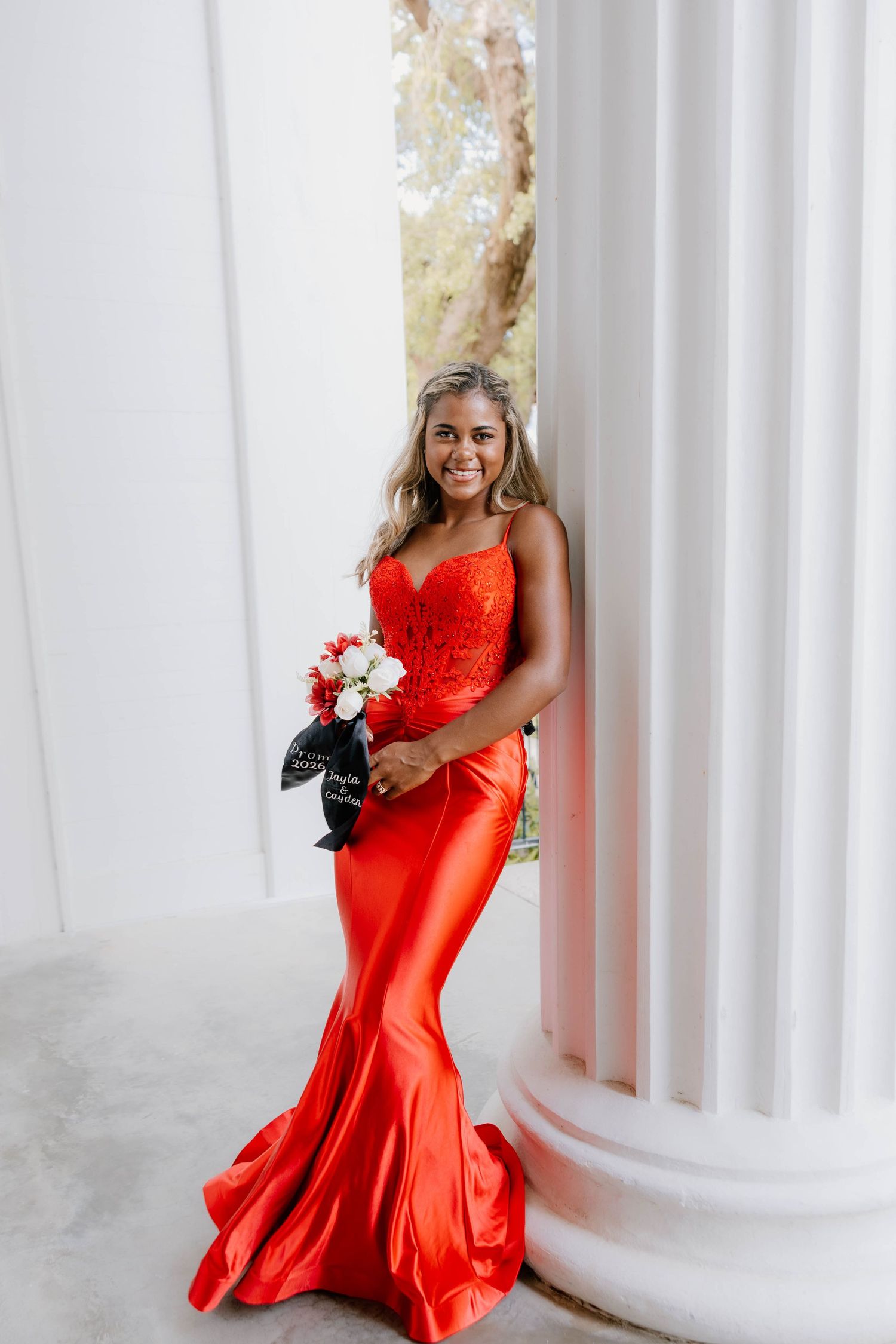 Young woman in elegant red mermaid prom dress holding bouquet, posing by white columns.